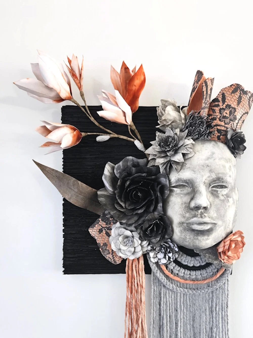 Mixed media art piece featuring a human face sculpture with flowers, leaves, and fabric elements arranged around it.