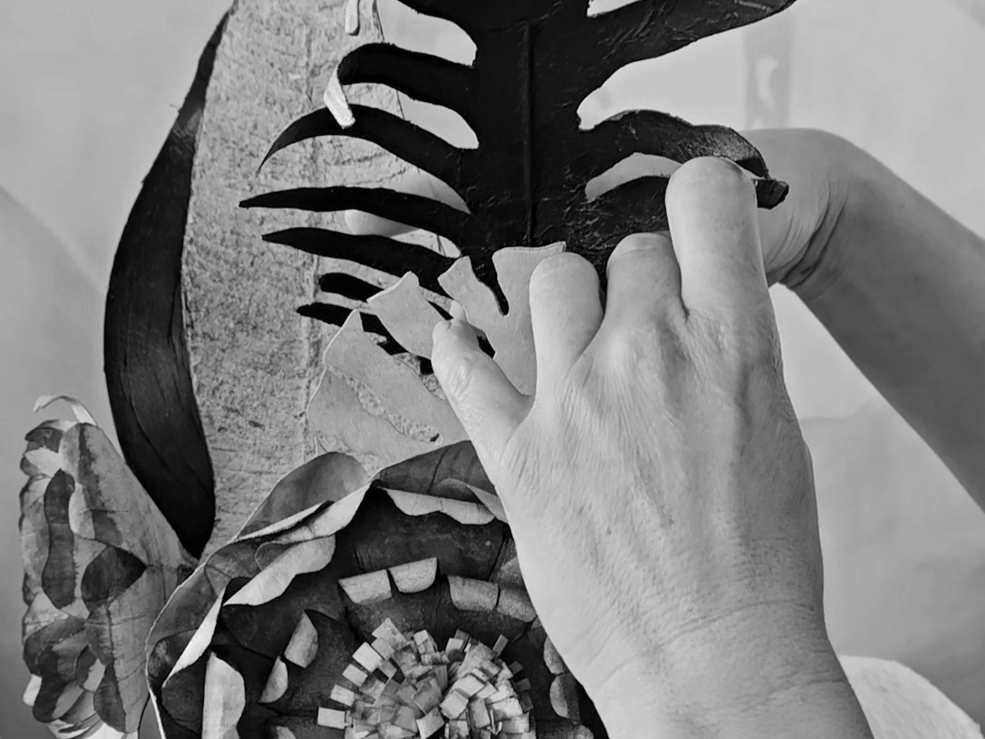 A person's hand is holding a cut-out paper leaf, with a stylized design that features floral patterns, and organic shapes. The background shows a textured surface.