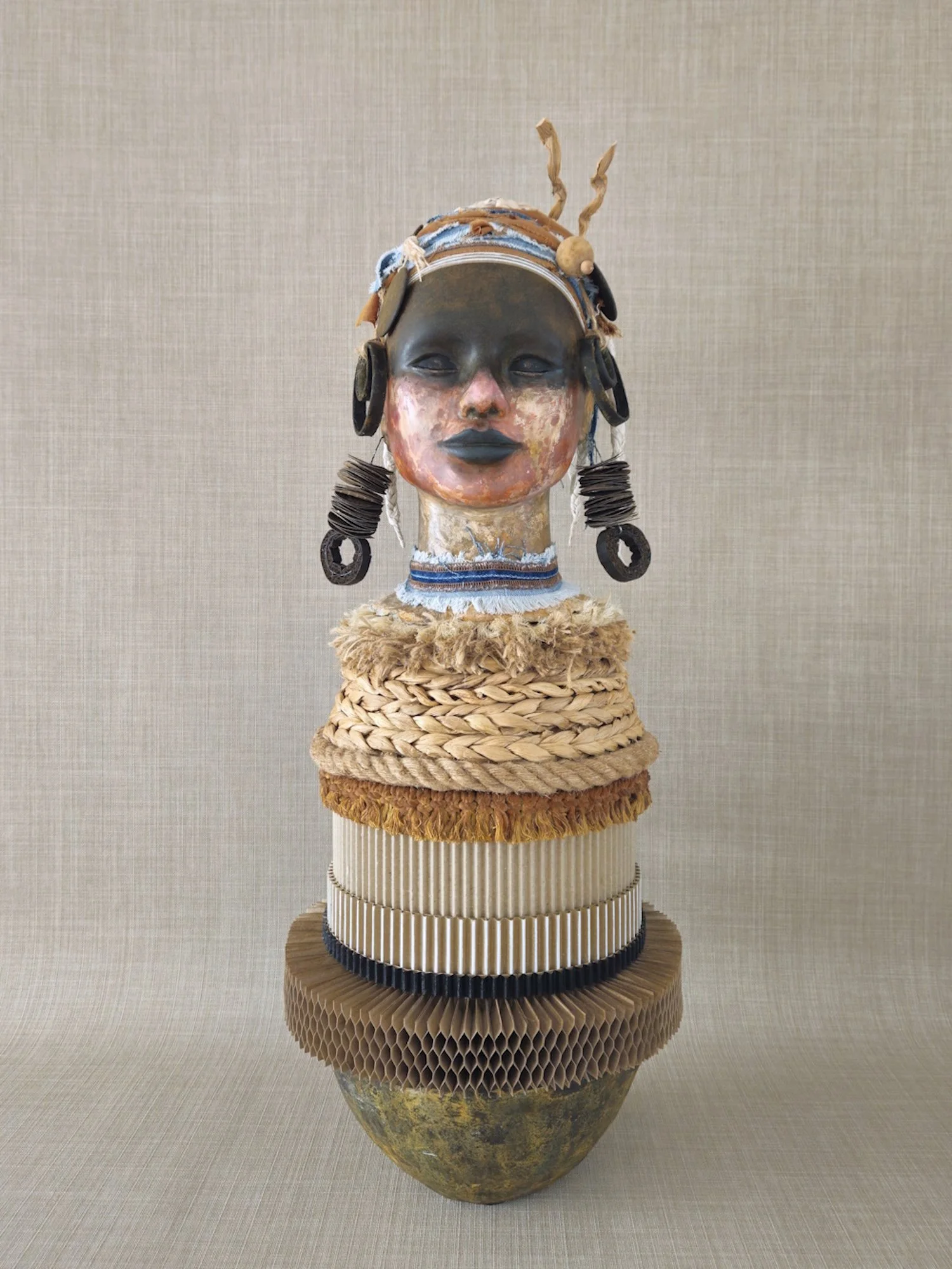 A sculpture of a woman's face with dark skin and closed eyes, adorned with earrings made of black and brown materials, and a headpiece with fabric, rope, and metallic elements. The face has a painted reddish-orange hue on the cheeks and chin, and the lips are painted dark blue. The sculpture's neck is decorated with a blue and white fabric choker, and the base is made of woven, textured, natural materials. The sculpture is displayed on a layered, circular platform with various textured and patterned materials, against a neutral, textured background.