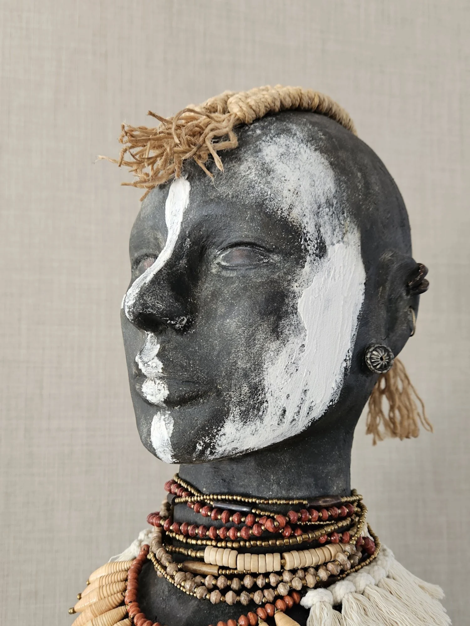 A black and white tribal mask with painted facial features, adorned with multiple layers of beaded necklaces and a rope headband, and a third eye earring.