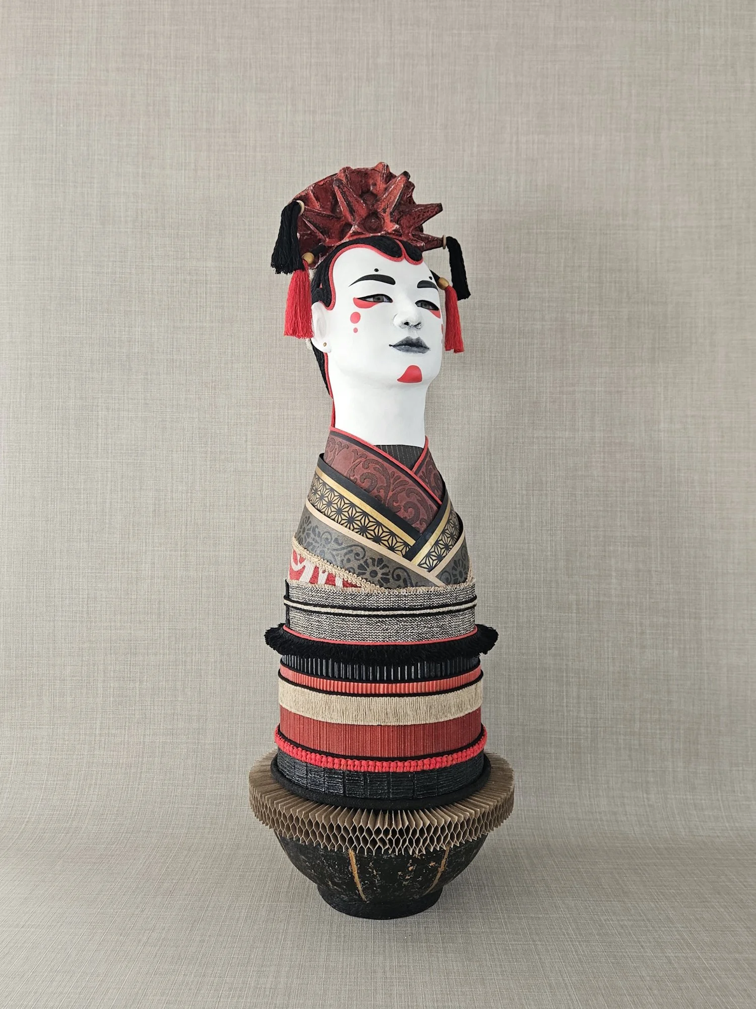 A Asian inspired sculpture displayed on a stand, decorated with a woven textile collar, with red, black, and white colors and intricate designs.
