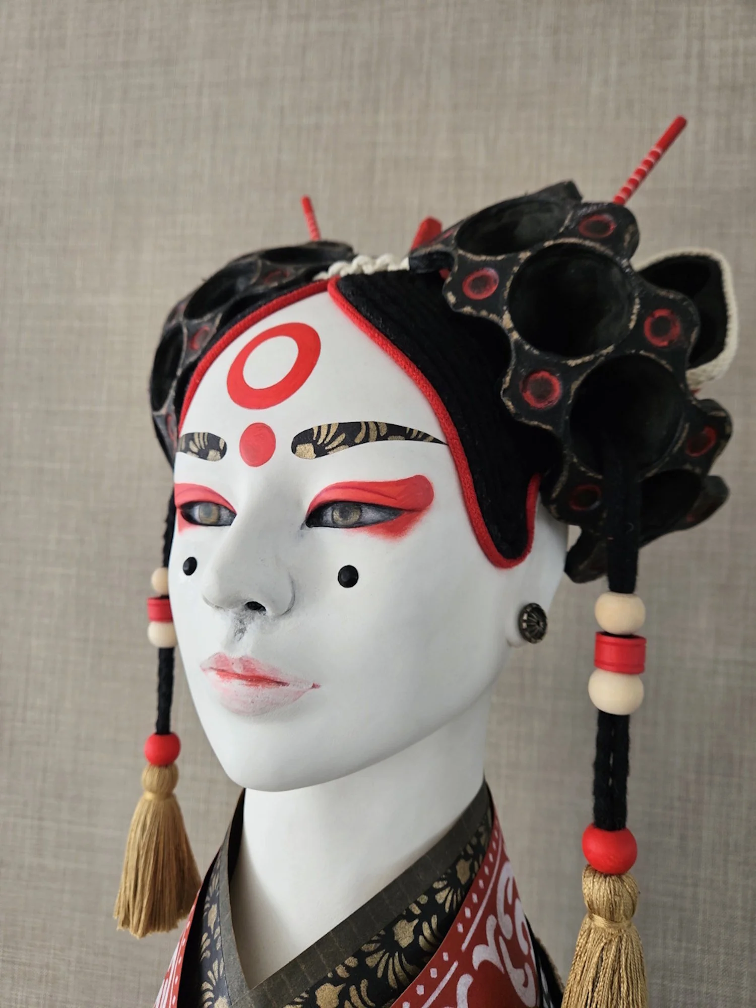 A  Japanese Noh inspired mask with intricate details and accessories, including beads and tassels, wearing a kimono with red and black patterns.
