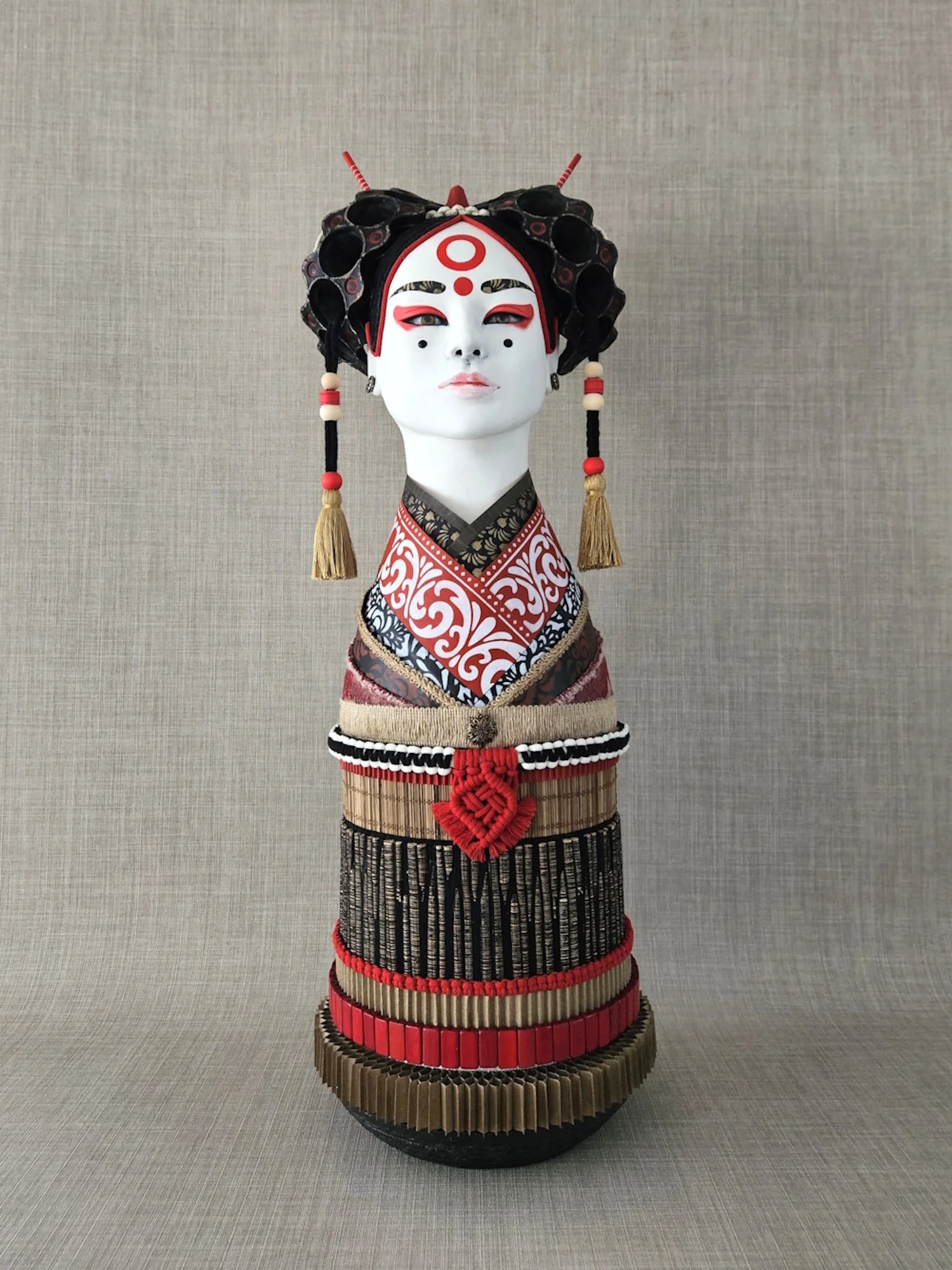 A decorative sculpture of a geisha with a traditional face mask, elaborate black headdress, and colorful layered clothing, standing on a round pedestal against a plain background.