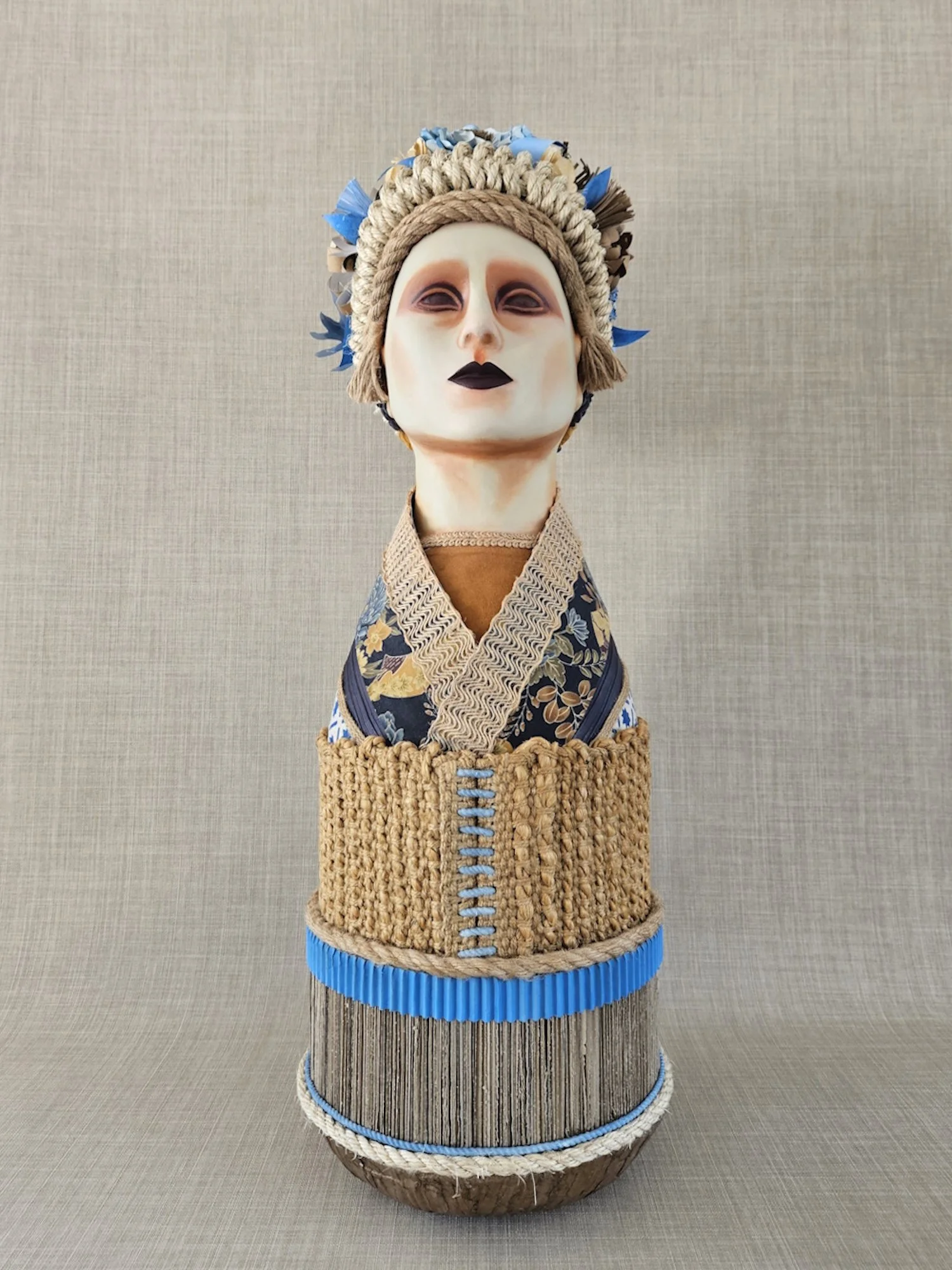 A figure with a painted face, black lips, and dark eyes, wearing a floral kimono and a woven basket-like torso, topped with a hat made of rope, straw, and paper, against a plain beige background.