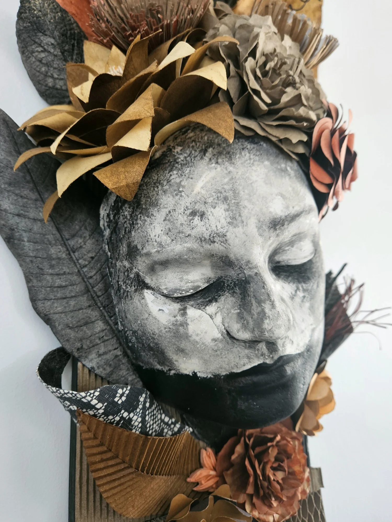 Close-up of a decorative gray stone face with a serene expression, adorned with a crown of various colored paper flowers, sitting on a wooden base.