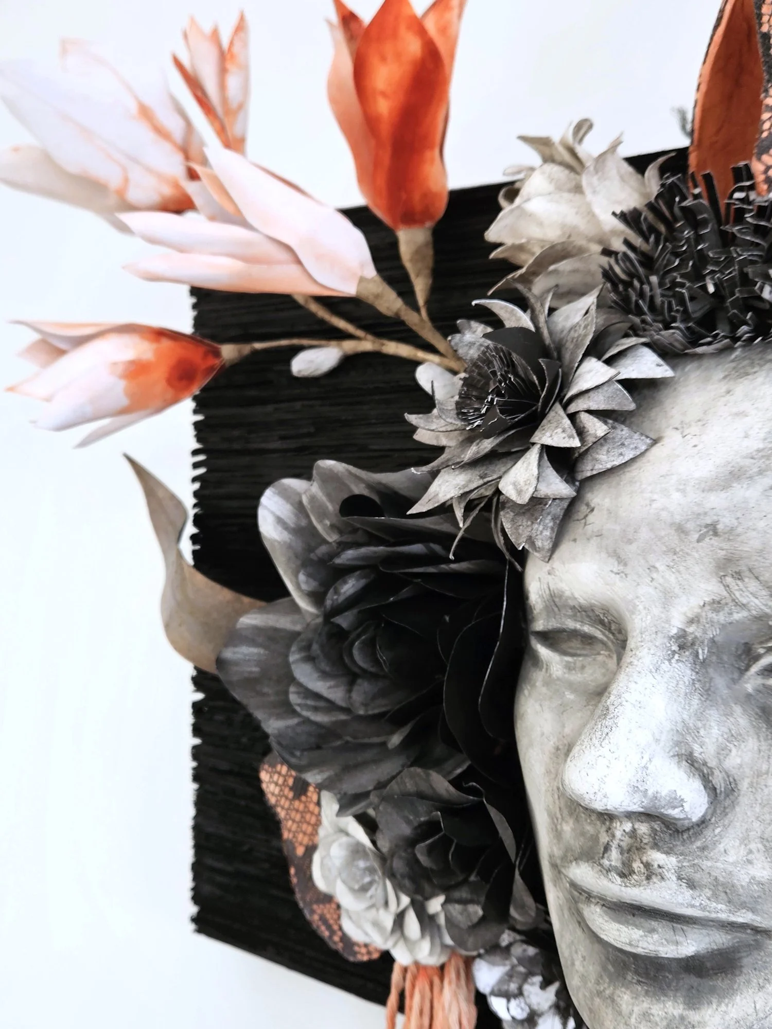 A sculpture of a human face with closed eyes, surrounded by paper flowers and foliage, and adorned with pink, orange, and cream-colored flowers.