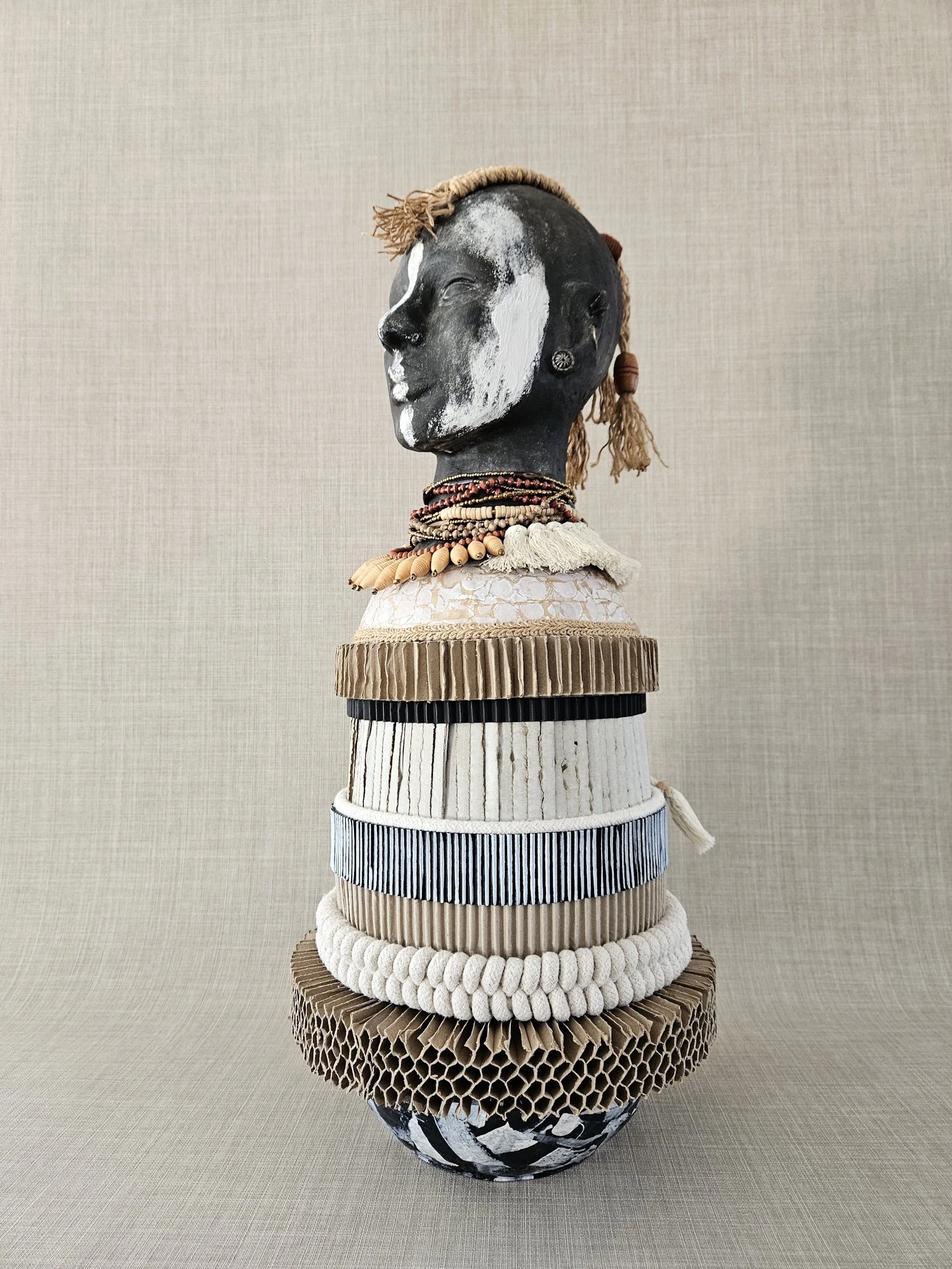 A sculptural art piece of a head with black and white painted face, decorated with multiple stacked rings made of various materials such as fabric, beads, and wood, set against a neutral background.