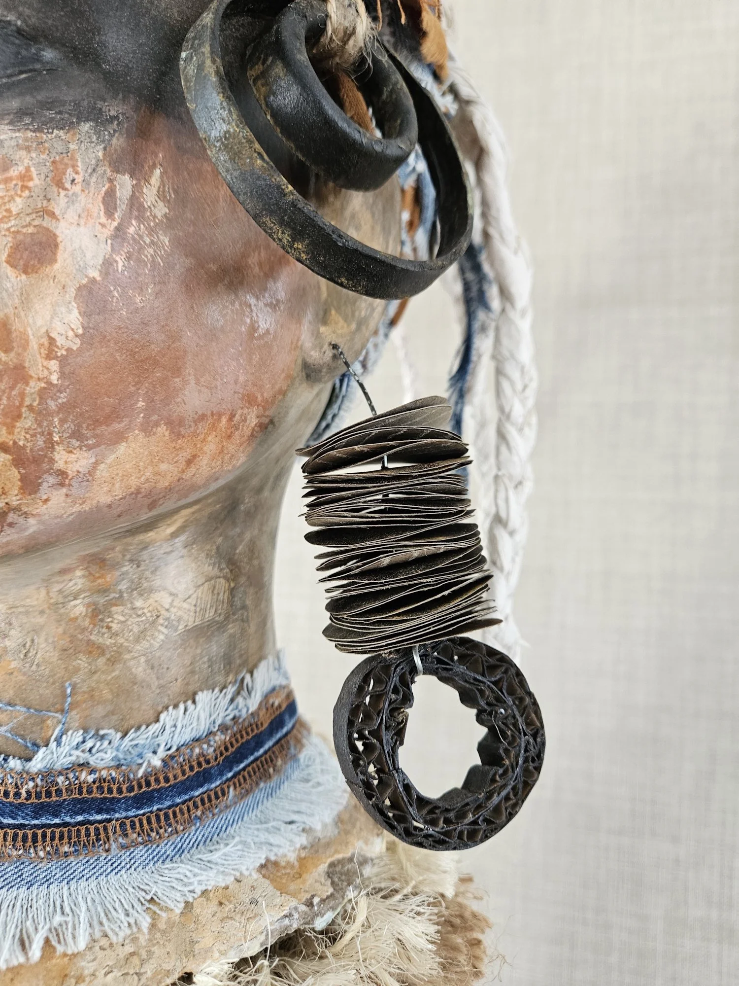 Close-up of a decorative hanging with black rings, fabric, and cardboard strips, displayed against a neutral background.