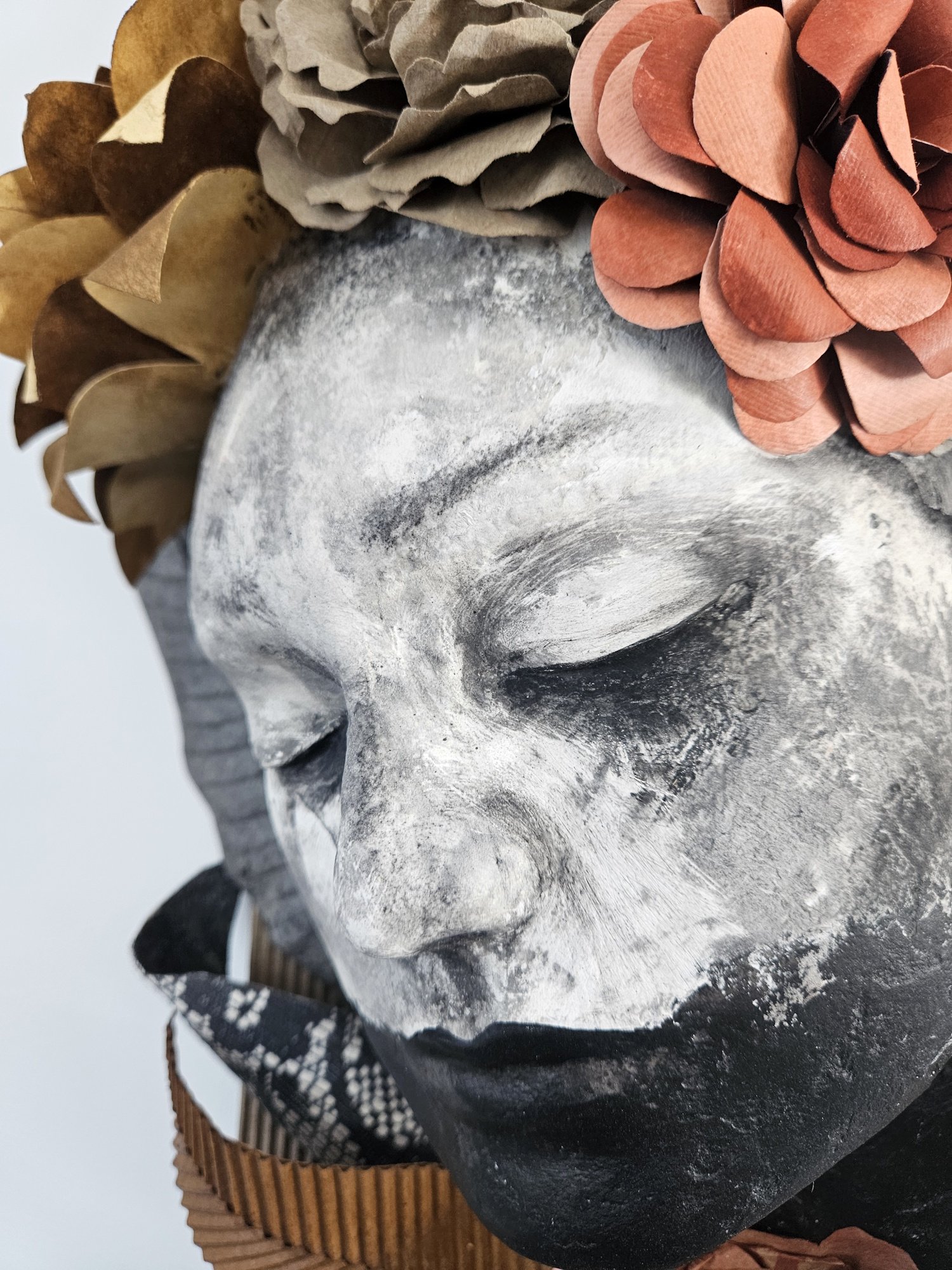 A black and white painted bust of a human head with wreaths of paper flowers in earthy tones resting on top.