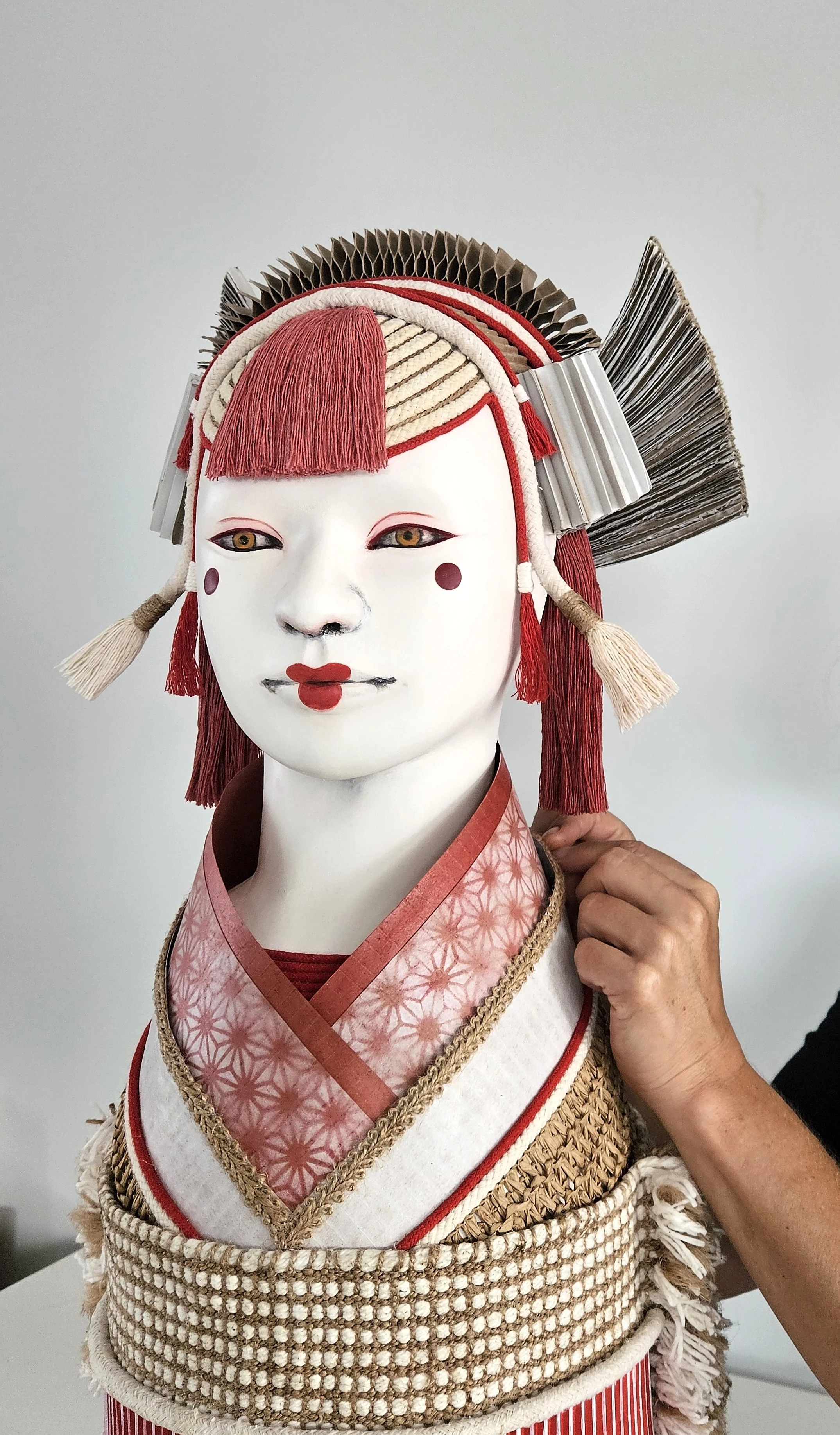 A Japanese inspired sculpture with a white face, red lipstick, and makeup, dressed in a pink kimono with floral patterns and elaborate accessories, including a headdress with paper and fabric elements.