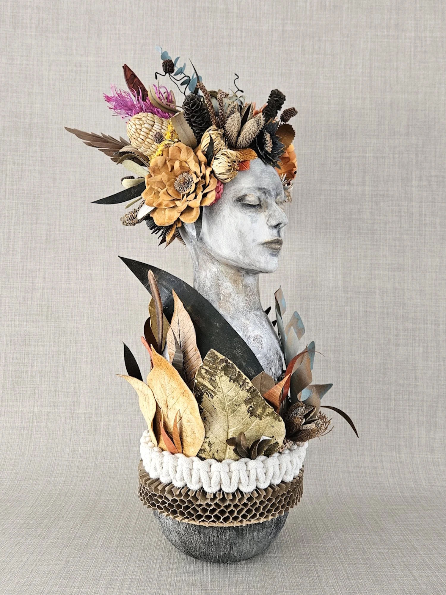 An art sculpture of a woman's face with closed eyes, featuring a floral and natural materials crown and accents, displayed on a textured surface against a plain background.