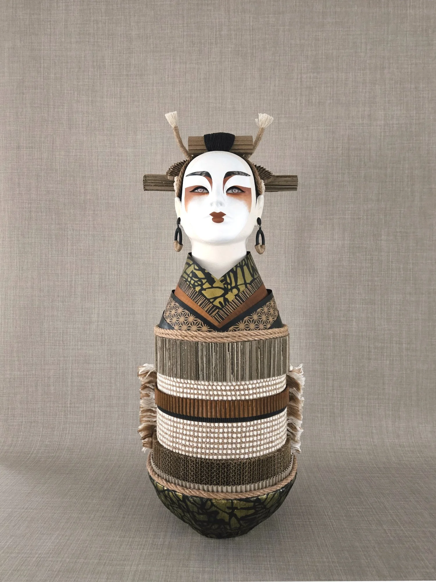A decorative art piece featuring a traditional Japanese mask with a serene expression, surrounded by layered woven and fabric elements, set against a plain neutral background.
