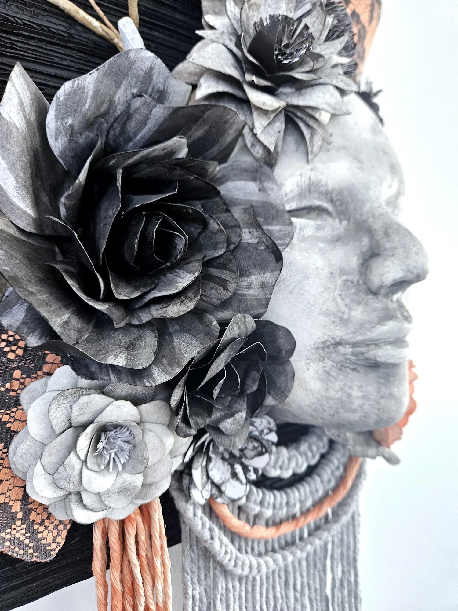 Close-up of a decorative face sculpture with black and gray rose-shaped embellishments and pink and gray braided accents on a textured background.