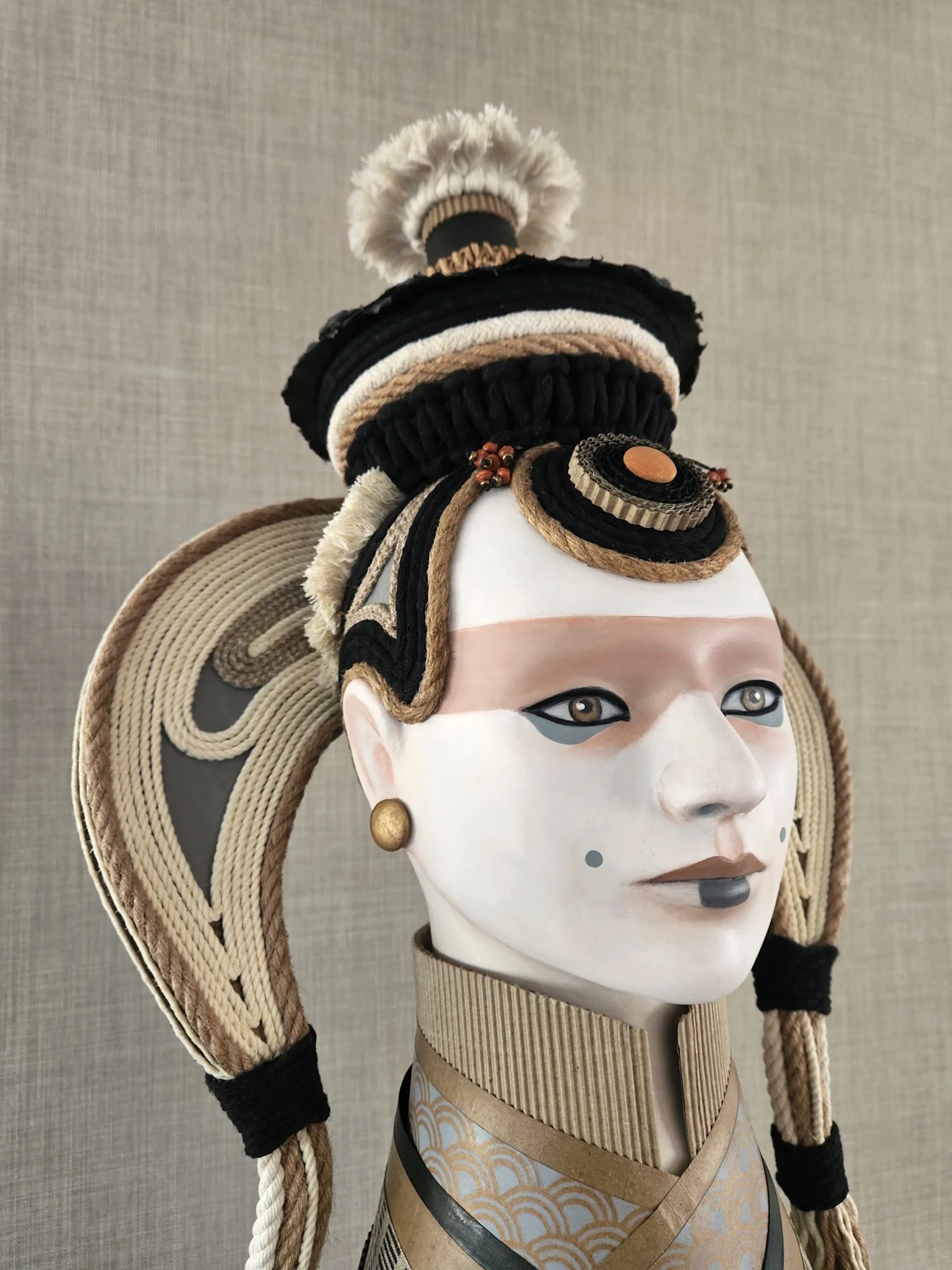 Traditional Japanese geisha mask with elaborate headdress including woven ropes, fabric, and decorative elements, set against a neutral background.
