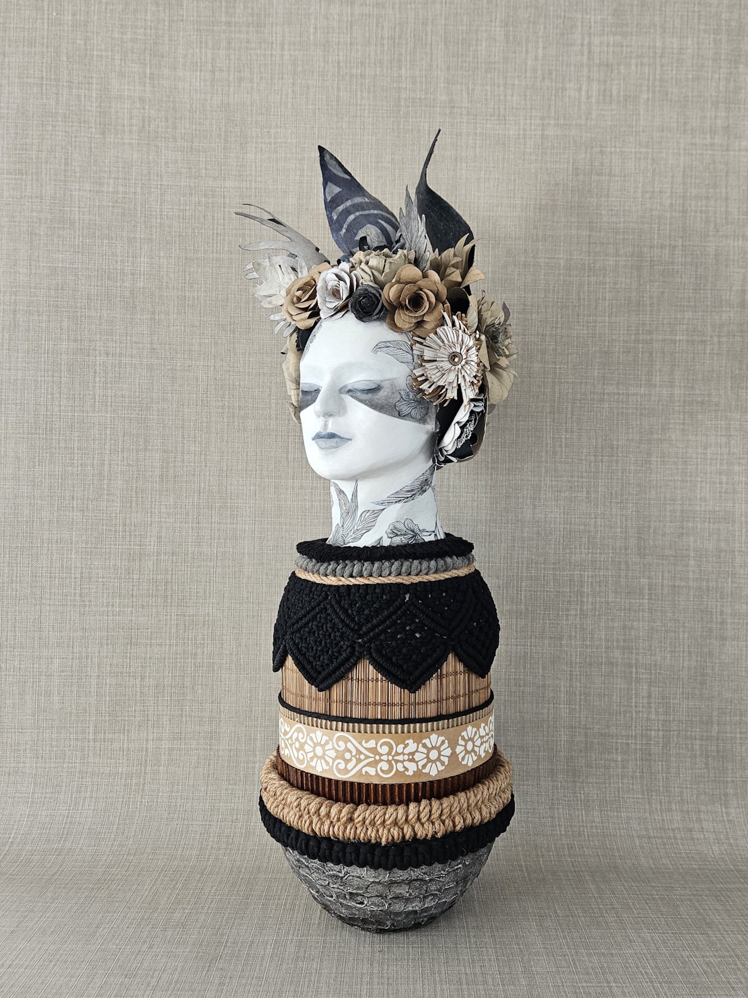 A sculpture with a painted face and closed eyes, adorned with a floral headdress made of paper, displayed on a series of stacked decorative rings with different textures and patterns.