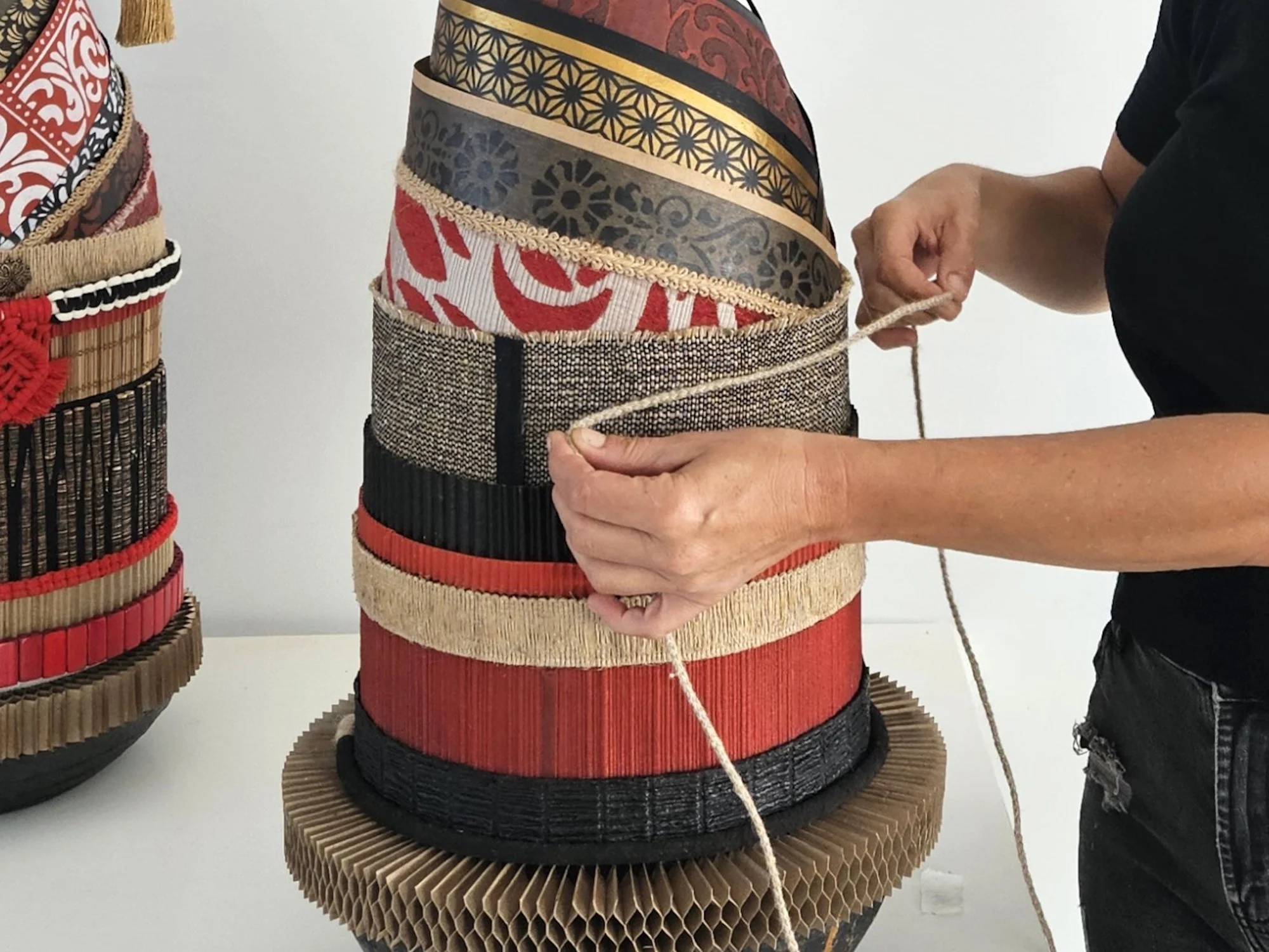 Artist holding a roped object made of layered, patterned materials including fabric, wood, and possibly paper, displayed on a stand with similar layered cylindrical objects.