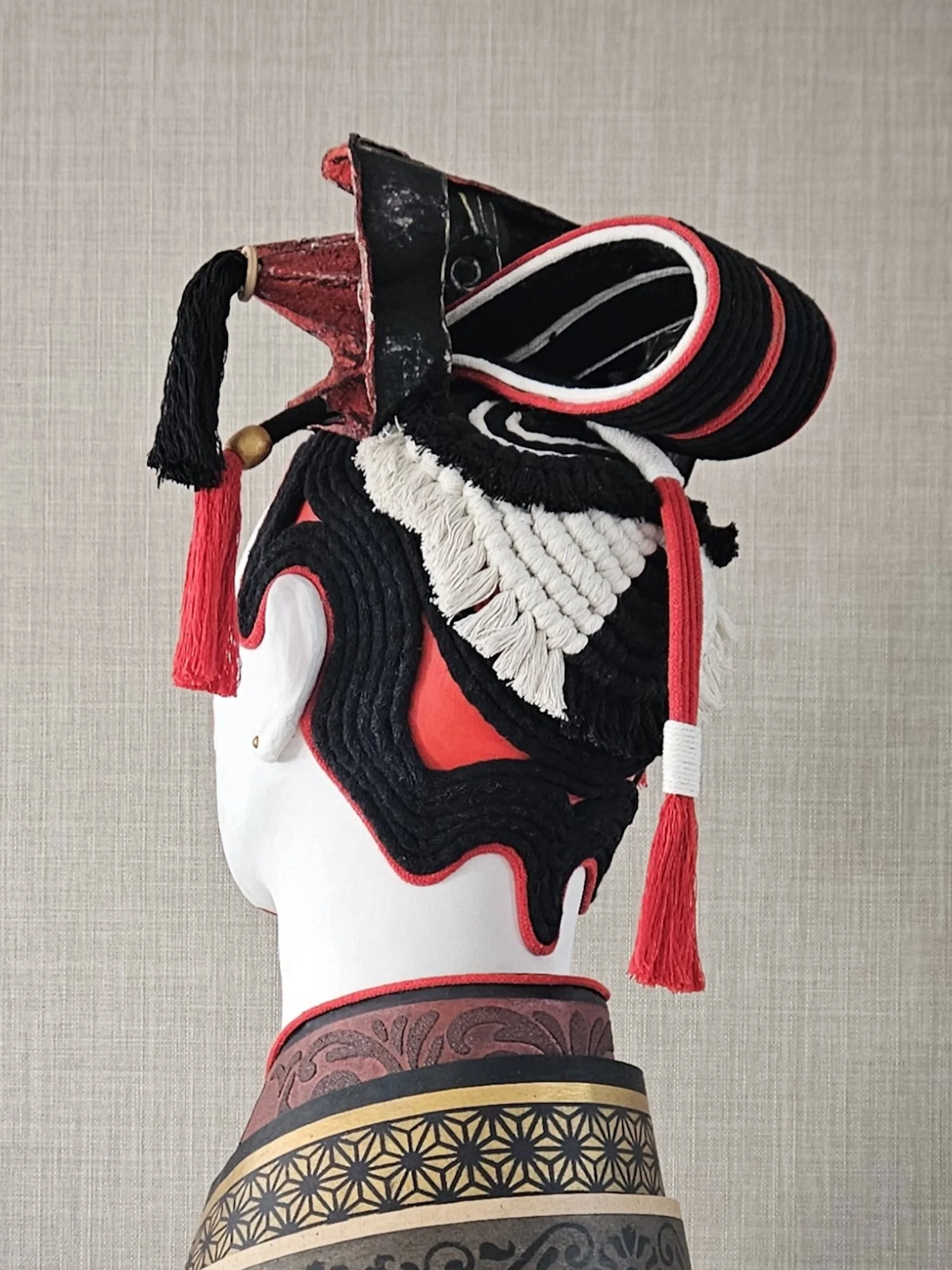 Tengu mask tilted sideways with red, black, and white colors, featuring a long nose, large eyes, and a black tassel hanging from the side, mounted on a display stand against a neutral background.