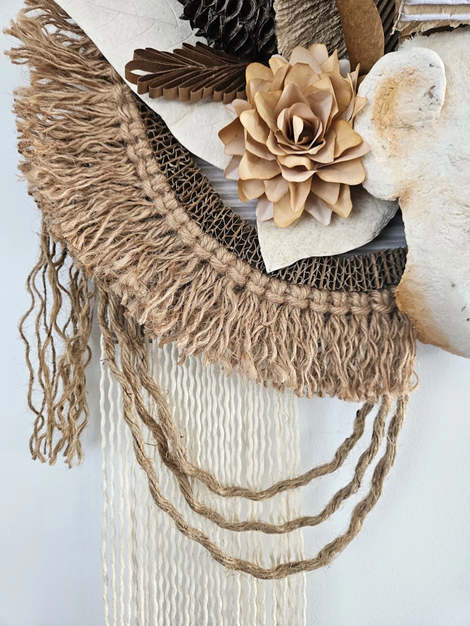Wall hanging made of natural materials including rope, wood, fabric, a stone, and paper flowers arranged in a layered design.