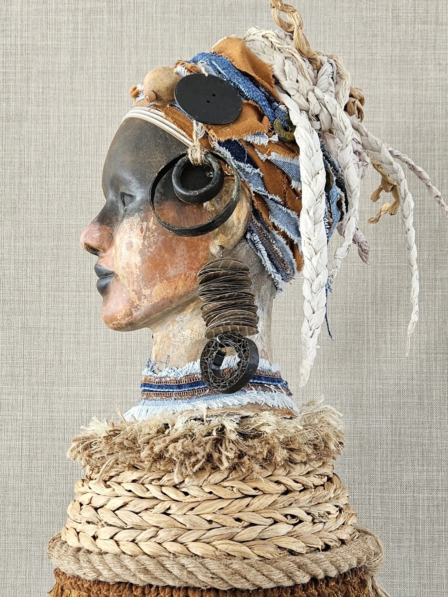 A colorful African tribal sculpture of a woman wearing jewelry made from natural materials, with braided hair, crafted from fabric, shells, and carved wood, mounted on a woven base.