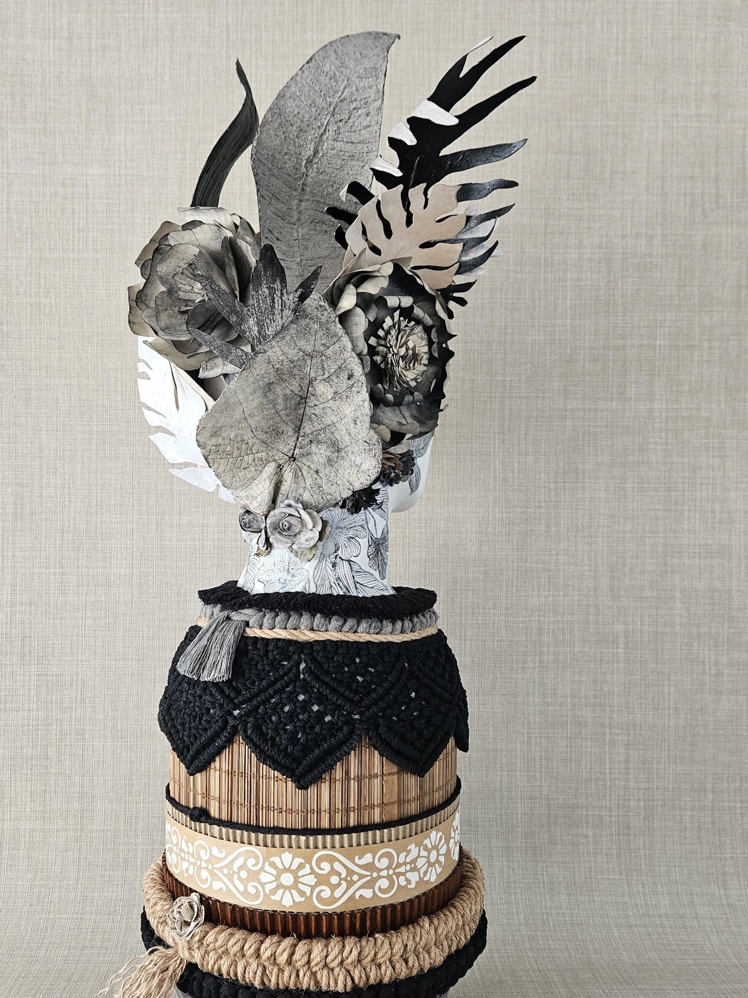 A multi-layered decorative arrangement featuring various natural and handmade materials, including leaves, flowers, and woven items, displayed on a stand against a plain background.