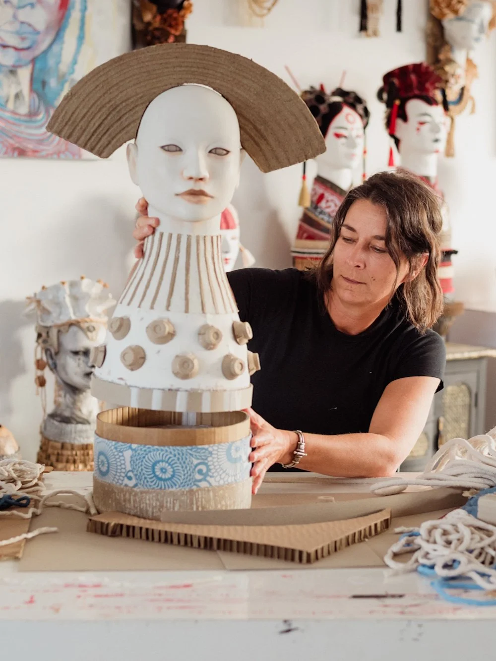 Artist assembling a sculpture in an art studio filled with similar sculptures and masks.