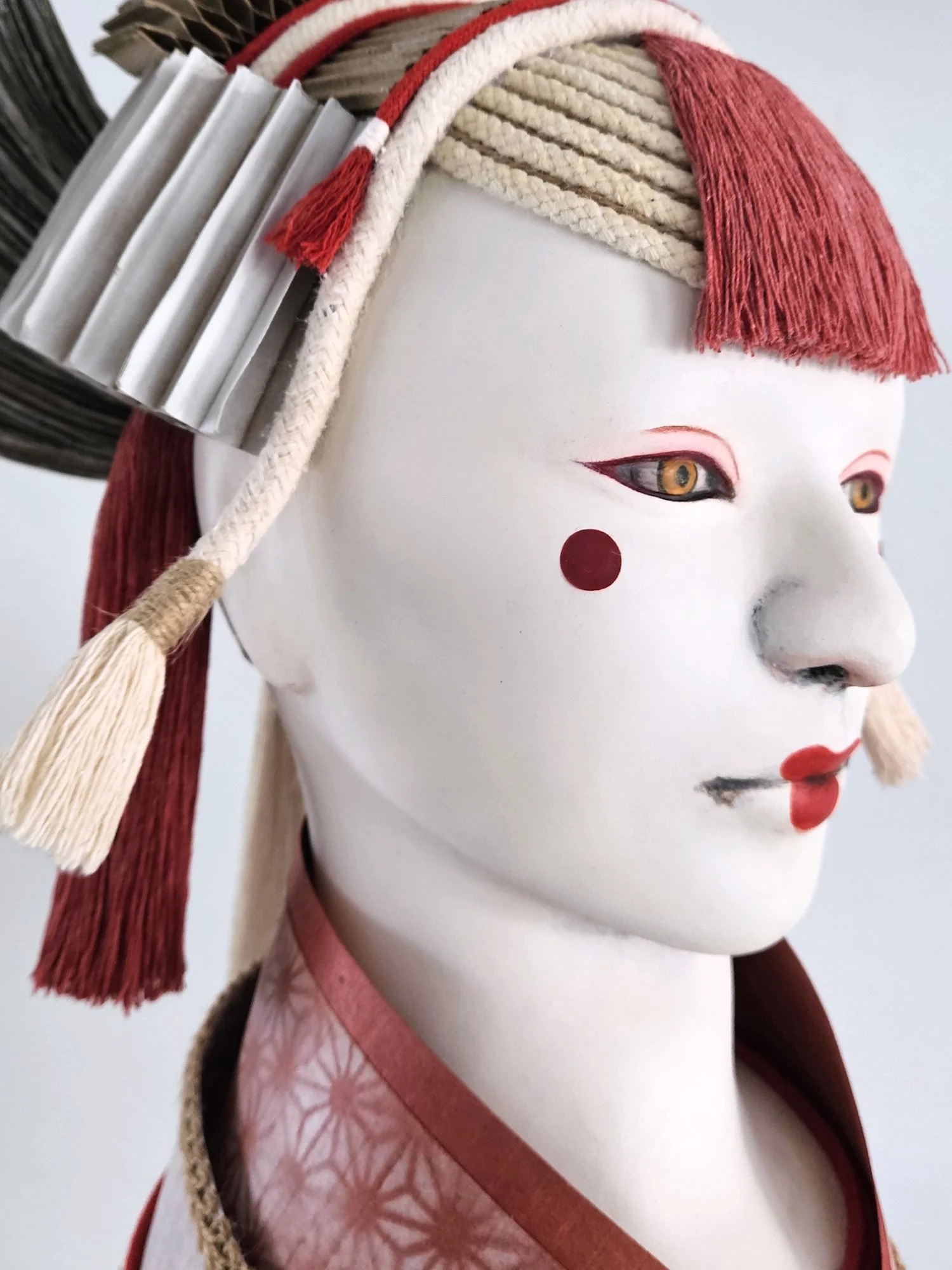 A Noh inspired mask with red, white, and black details, featuring a white face, red dots on the cheeks, and makeup around the eyes, with elaborate headgear decorated with rope, tassels, and fabric.