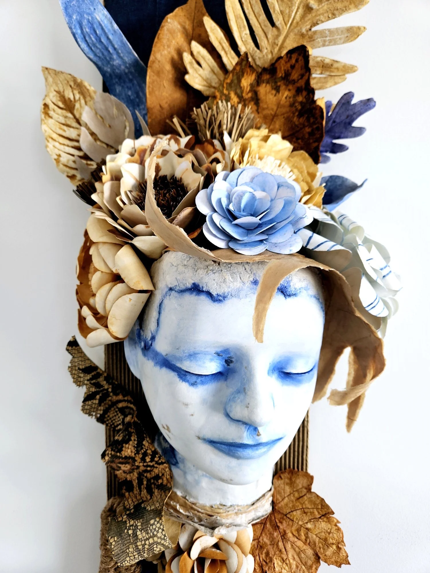 An artistic sculpture of a human face with closed eyes, painted white with blue accents, decorated with dried leaves, flowers, and feathers arranged on the head.