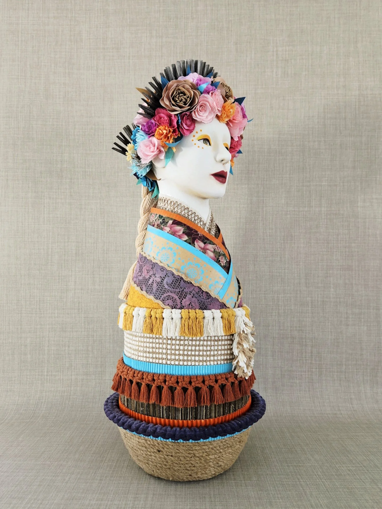 A sculpture of a woman with a paper flowers  headpiece from Slavic inspiration. The head is placed on a textured woven basket-style stand, with a neutral beige background.