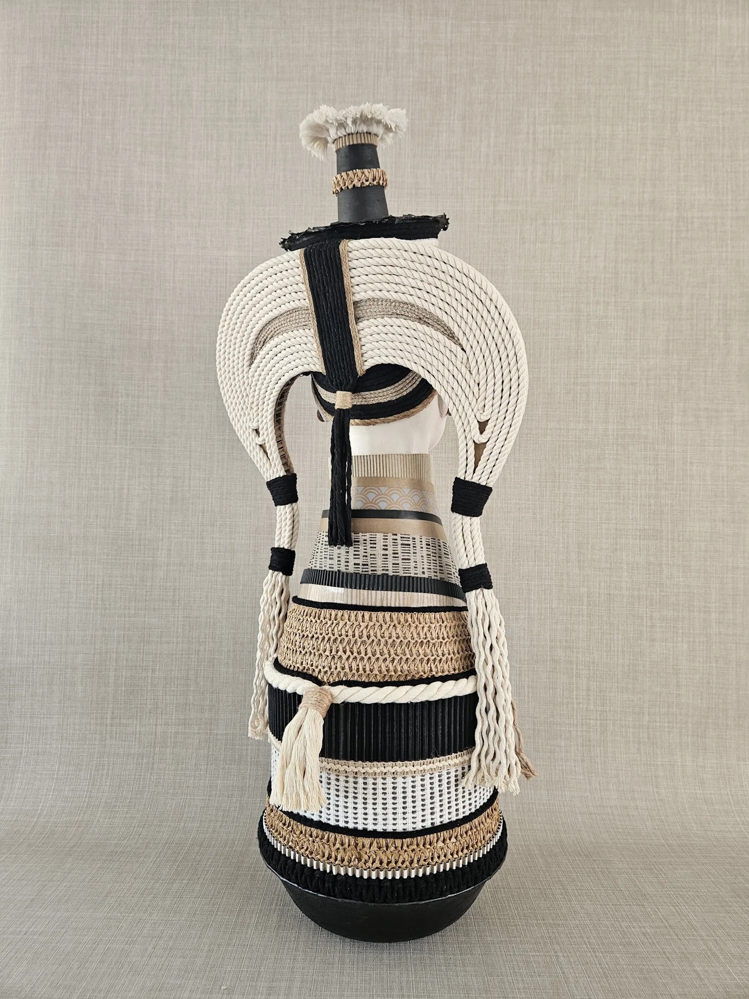 Sculpture of a human figure with a body made of woven and textured materials, wearing a tall hat with layered rings, against a plain, textured background.