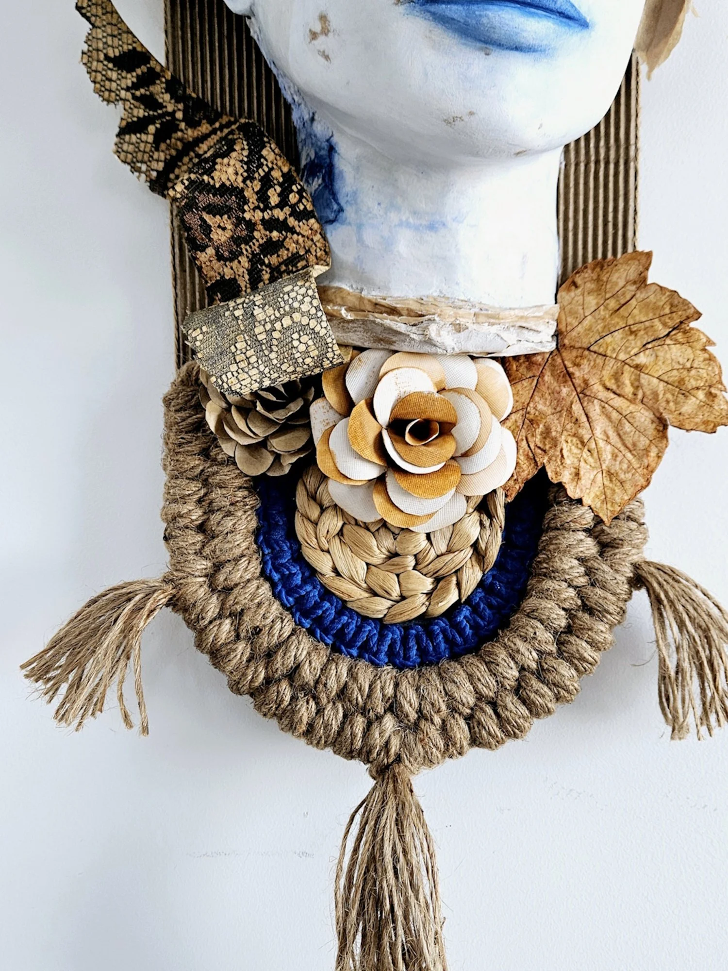 Decorative wall hanging with a bust sculpture, paper leaves, fabric flowers, and woven rope accents on a white background.