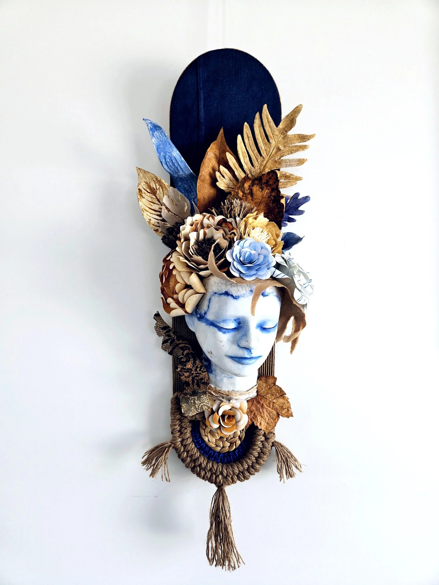 A decorative wall art piece featuring a white paper mache human face sculpture with closed eyes, adorned with various flowers, dried leaves, and feathers on the top and around the head, with a woven necklace at the bottom.