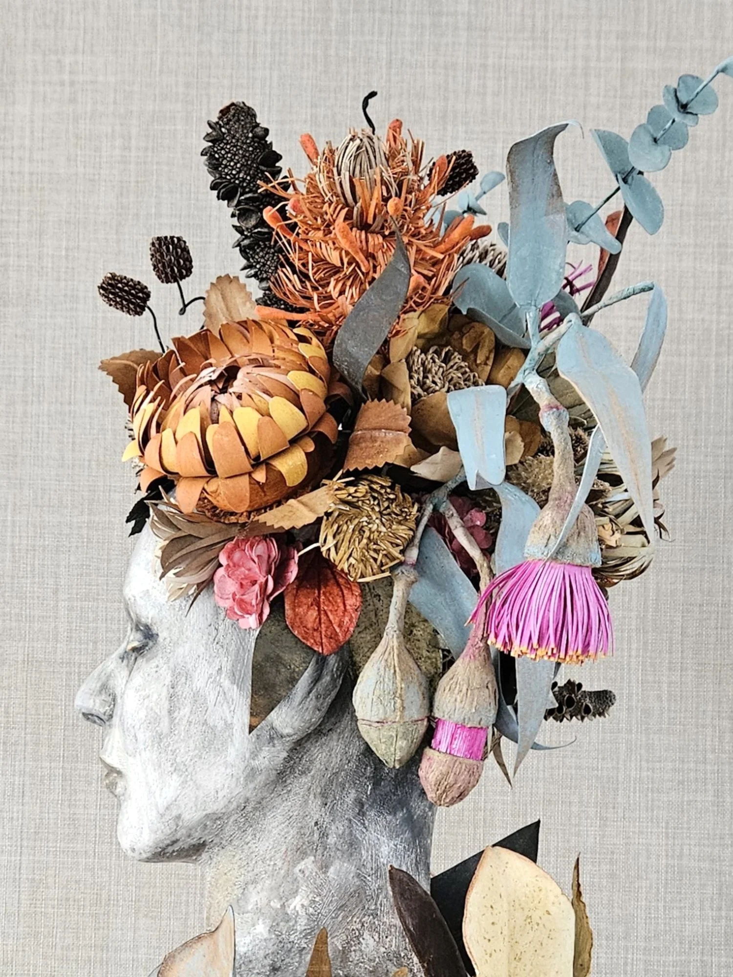 A mixed bouquet of paper flowers, leaves, and seed pods on a driftwood branch against a neutral background.