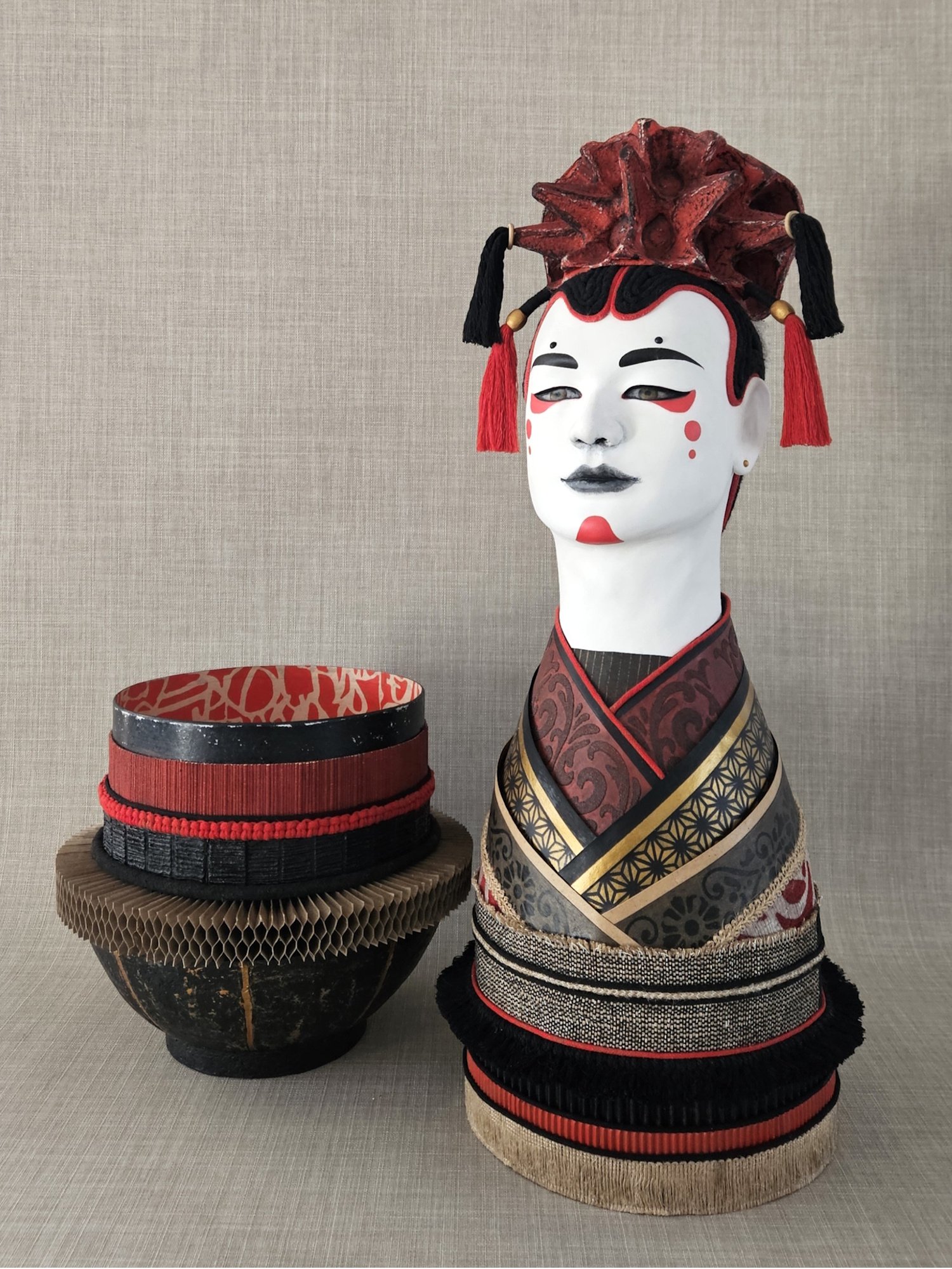 A head sculpture wearing a Japanese inspired headpiece and makeup, with a large red and black hair ornament. Next to the mannequin is a container decorated with intricate red and black patterns.