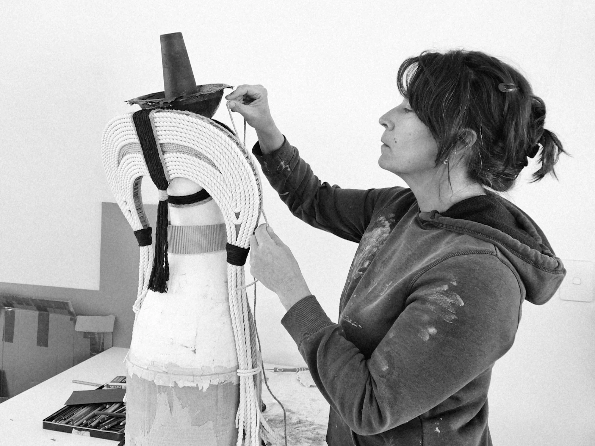 A woman artist working on a head sculpture. The sculpture is draped with white and black braided rope decorations. The woman is holding a string, adjusting the display.