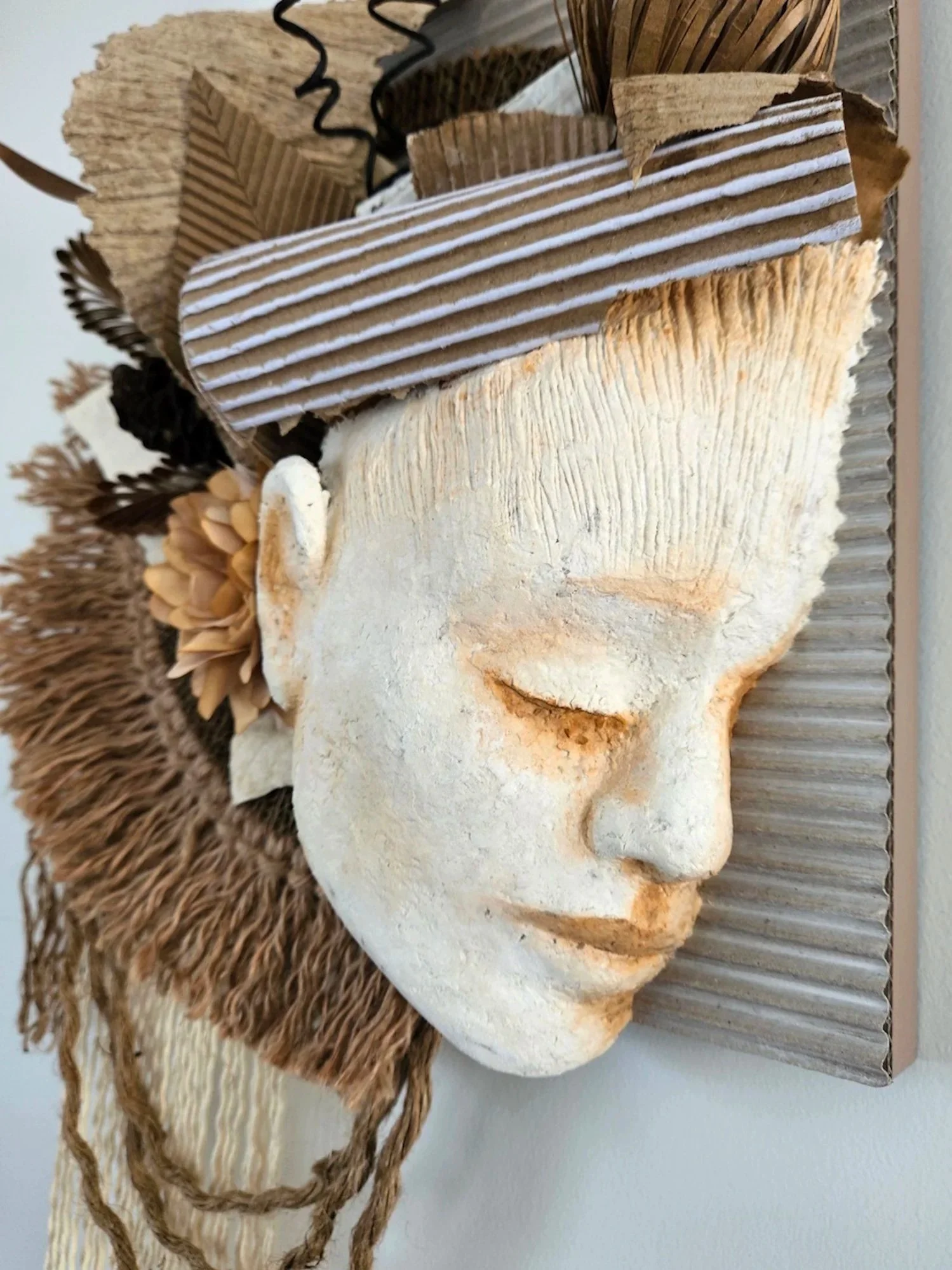 A decorative wall art piece featuring a carved white paper mache face with closed eyes and detailed features surrounded by paper flowers and foliage, textured paper, and natural elements arranged on a striped wooden board.