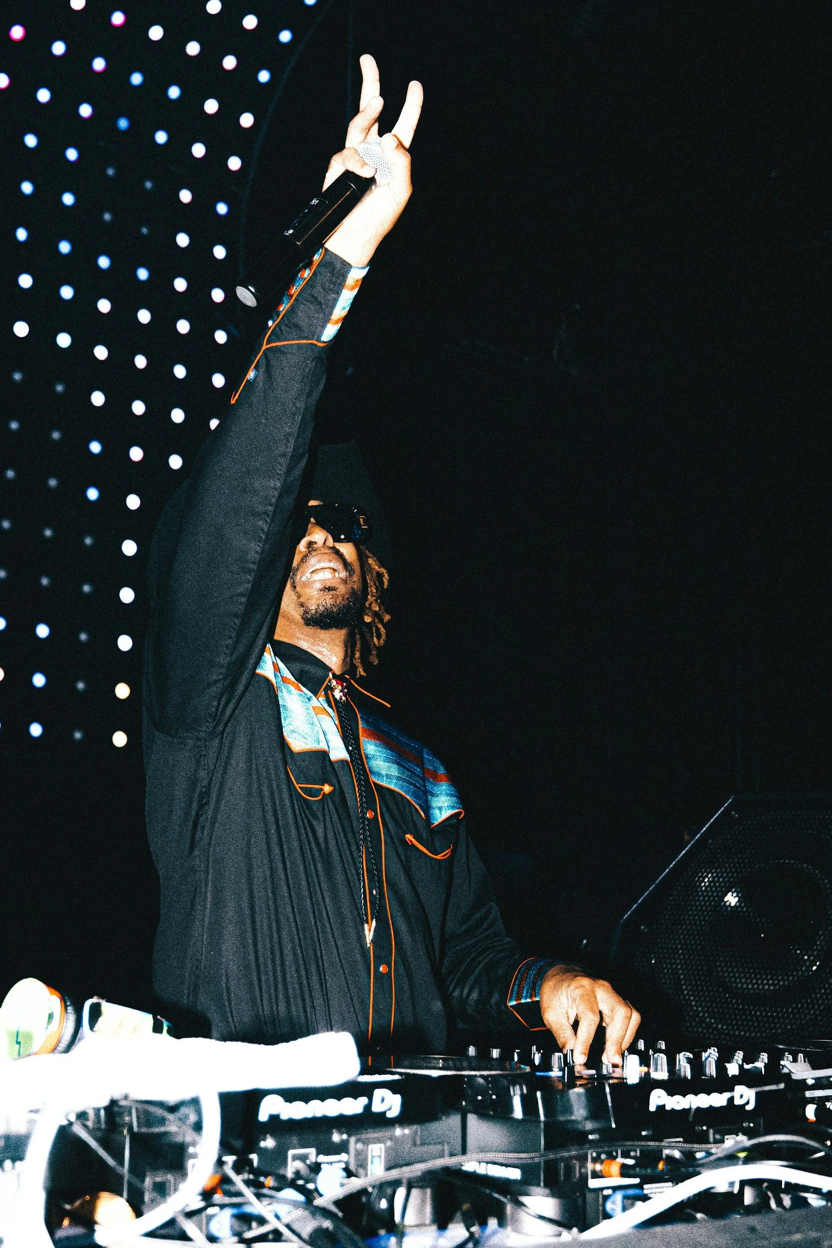 A male DJ with dreadlocks and sunglasses playing music at a dark venue, with a background of black perforated panels with blue and purple lights.