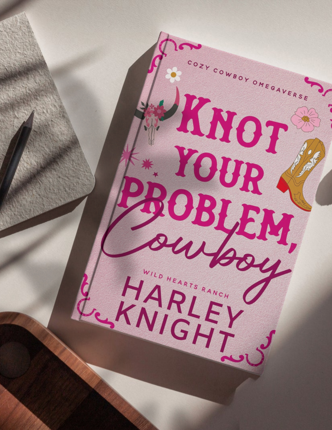 Book titled 'Knock Your Problem, Cowgirl' by Harley Knight on a white surface with a shadow, adjacent to a piece of textured paper and a pen, with a decorative western boot illustration on the cover.