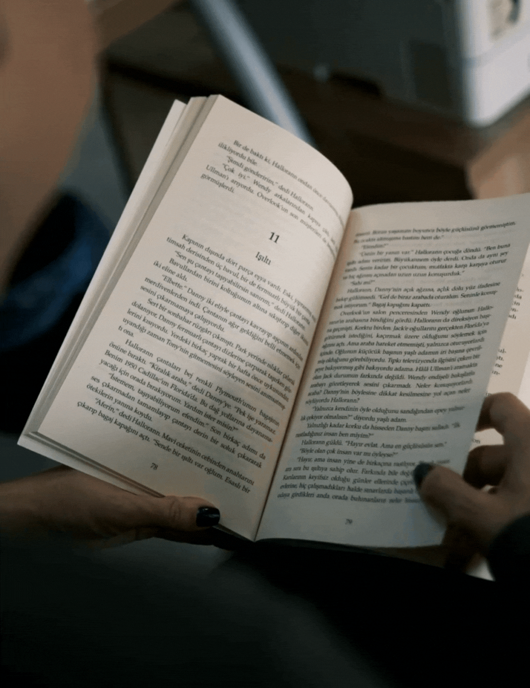 Person holding an open book written in Turkish, showing pages 10 and 11, with a blurry background of a table or desk.