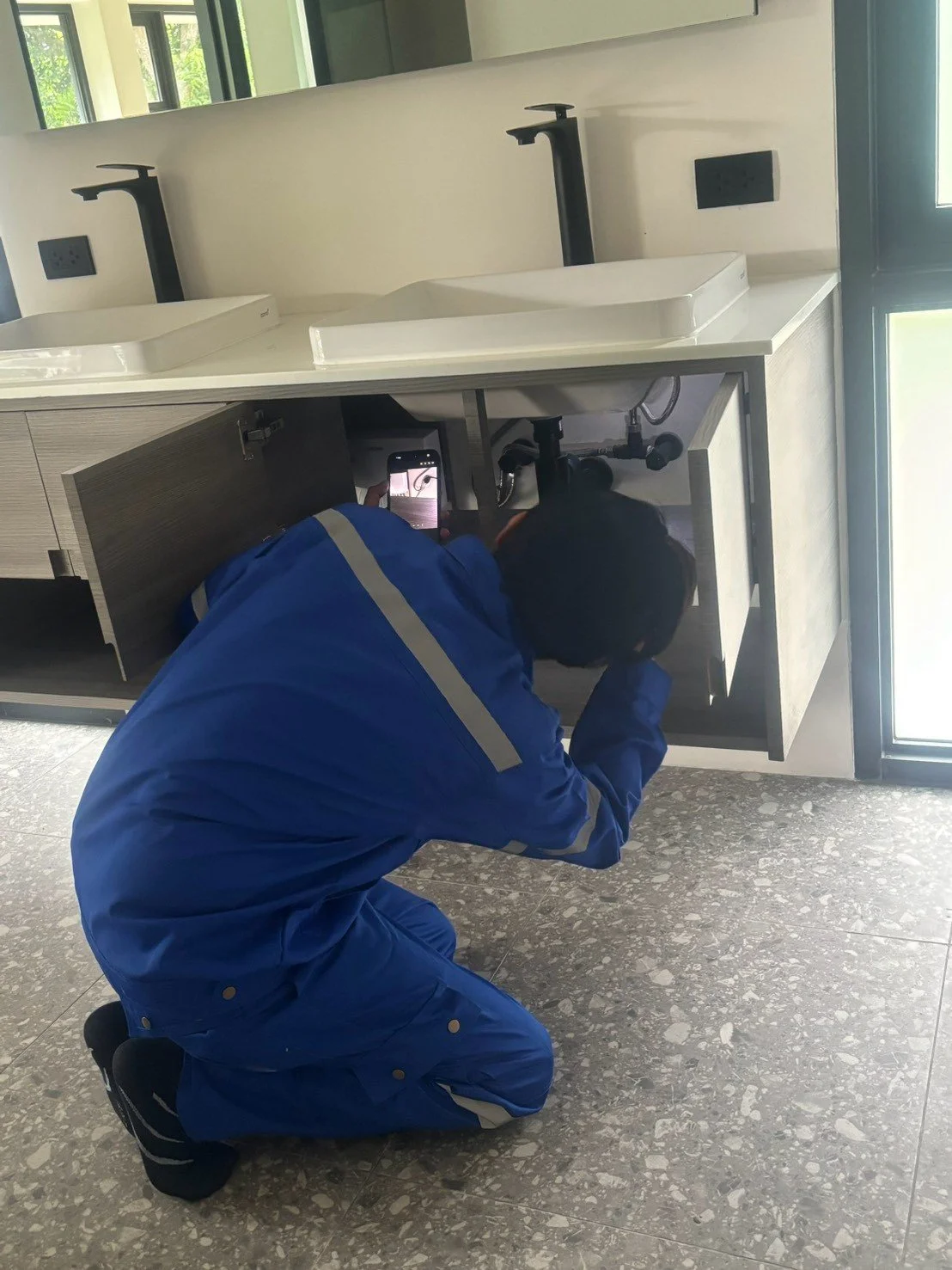 A person in a blue uniform kneeling under a bathroom sink, taking a photo with a smartphone.