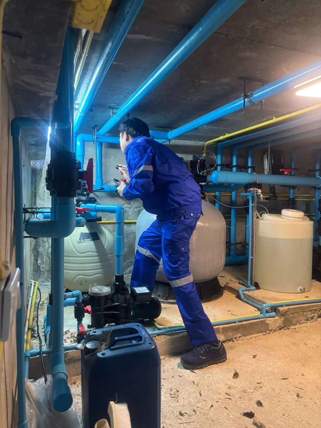 A worker in blue overalls inspecting or adjusting plumbing and filtration system in a basement or utility room.