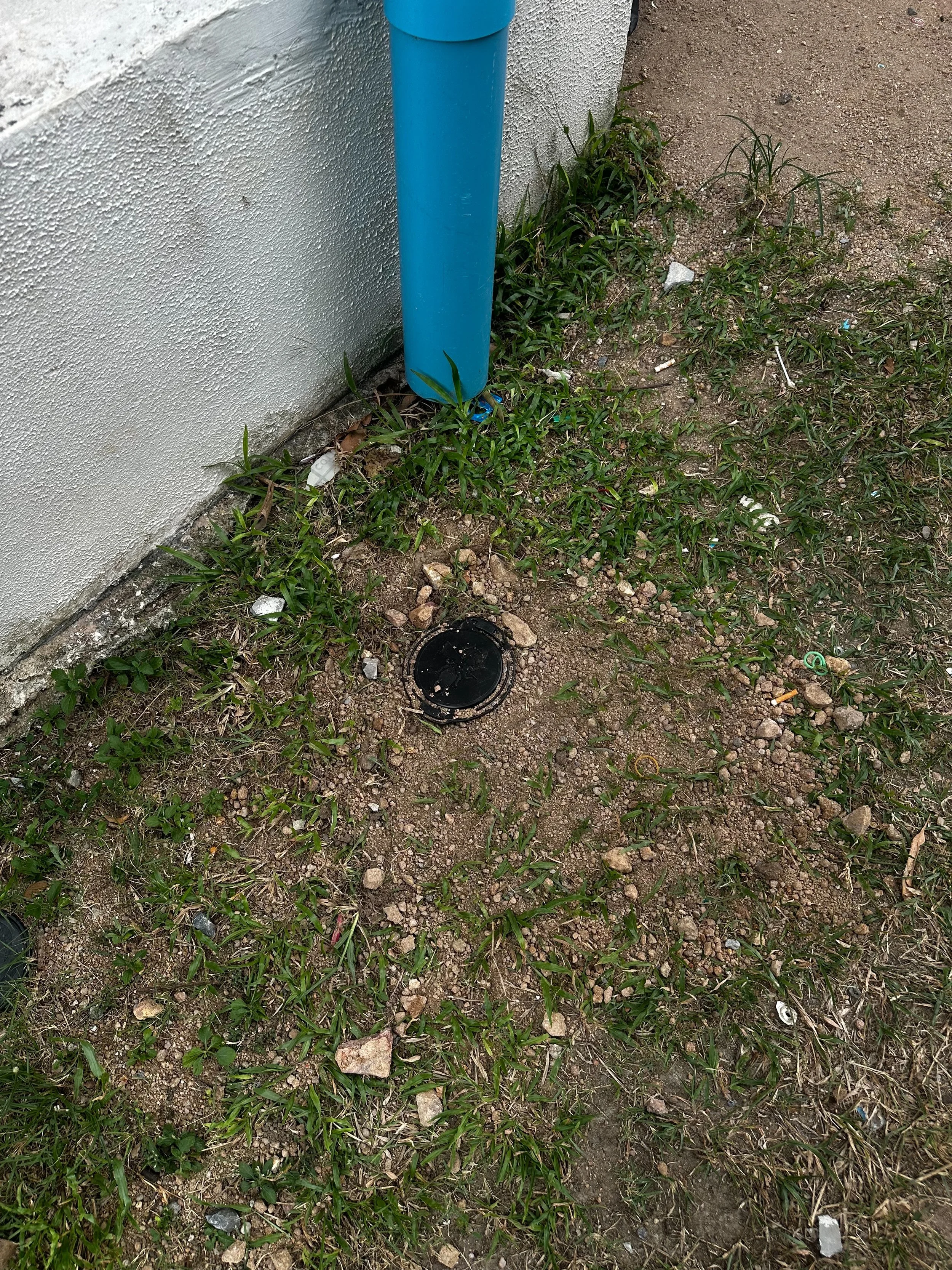 A blue cylindrical object attached to a white exterior wall, with grass, dirt, and small debris on the ground beneath it.