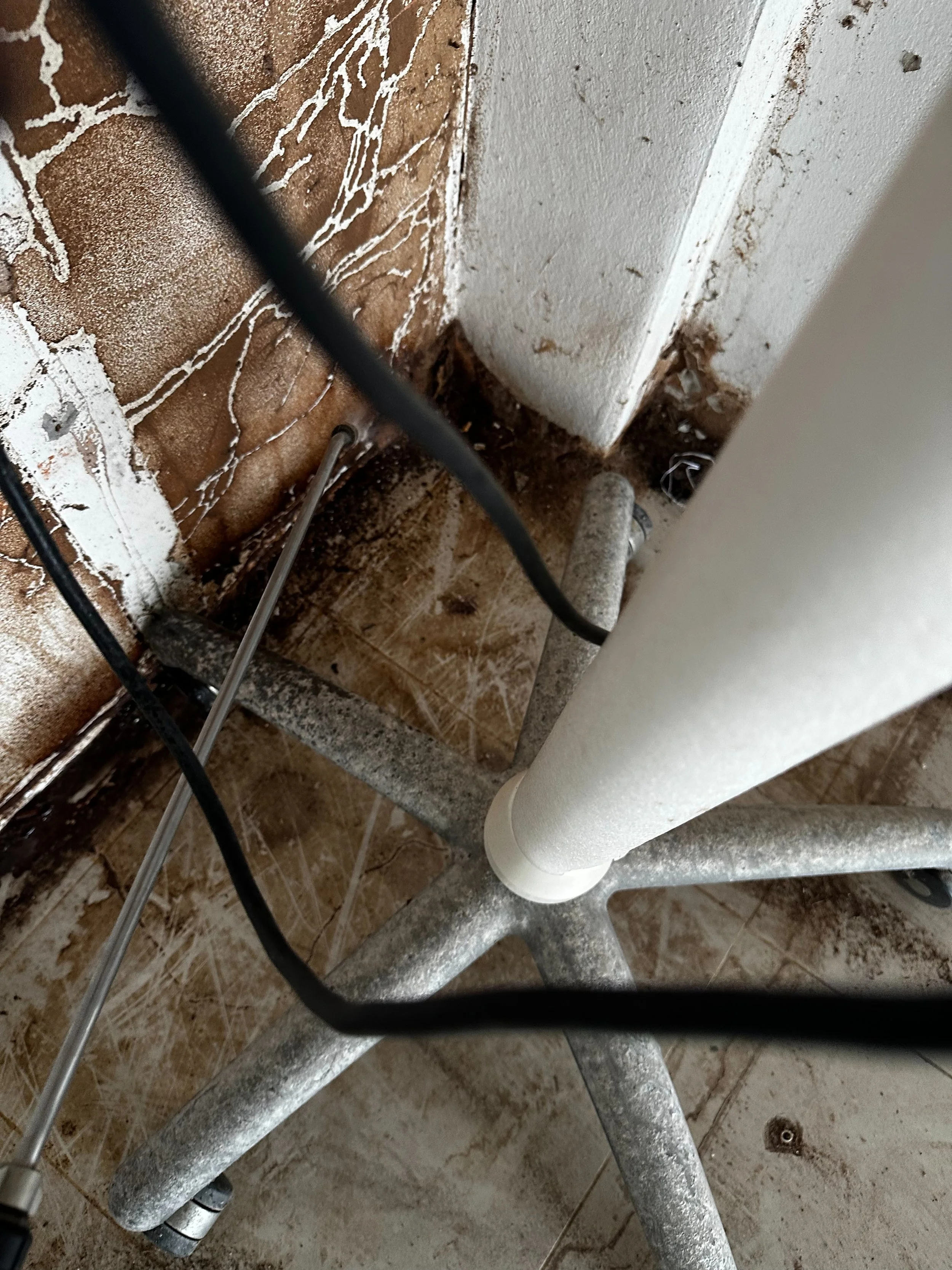 Close-up of a small section of a bathroom, showing parts of a white sink, pipes, and a rusty metal frame, with dirty walls and floor.