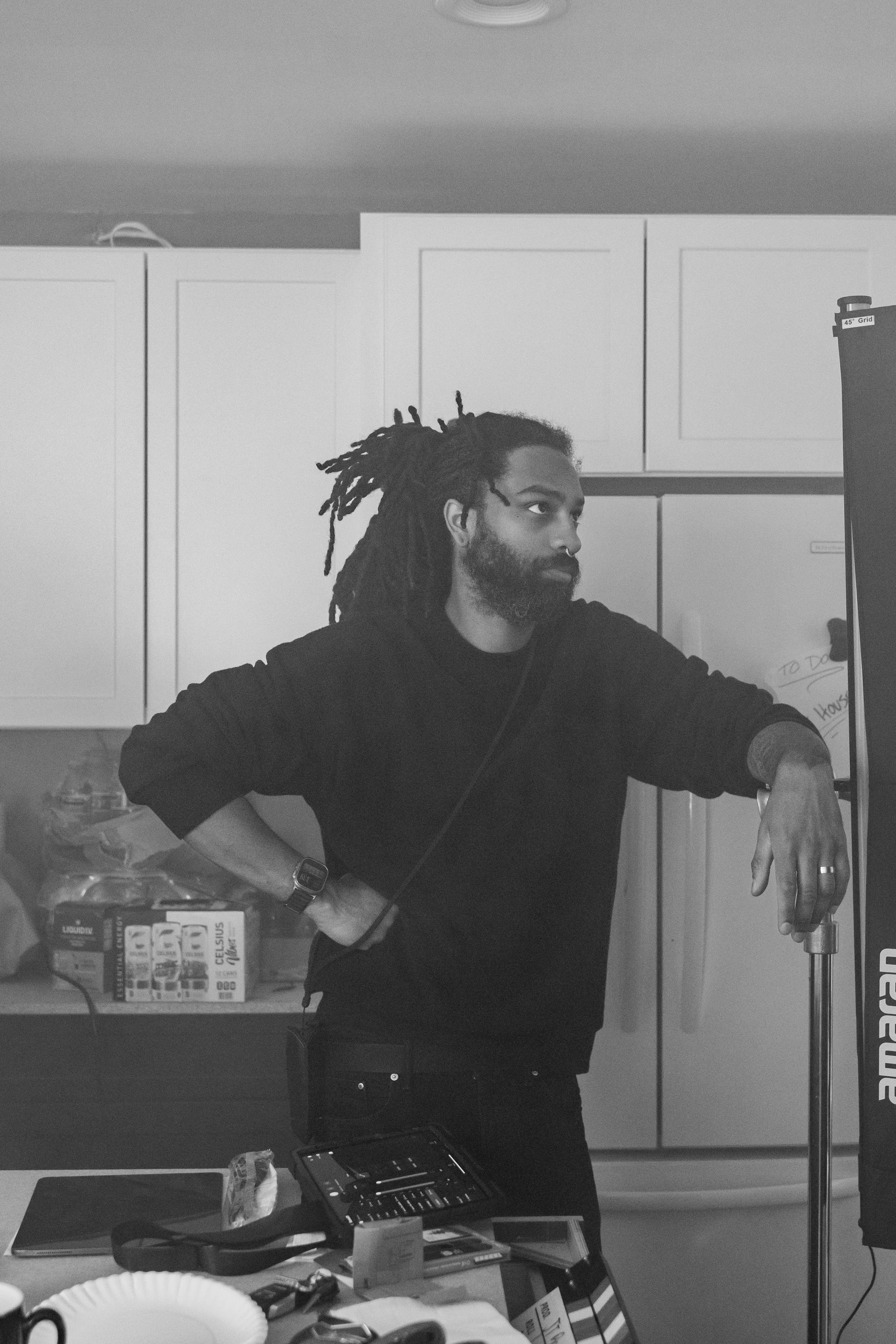 D'Andre poses on a commercial set, with his right arm resting on a stand. He is looking slightly upward and to his right. The background contains packaging boxes and various items on a table, with a whiteboard partially visible on the right.