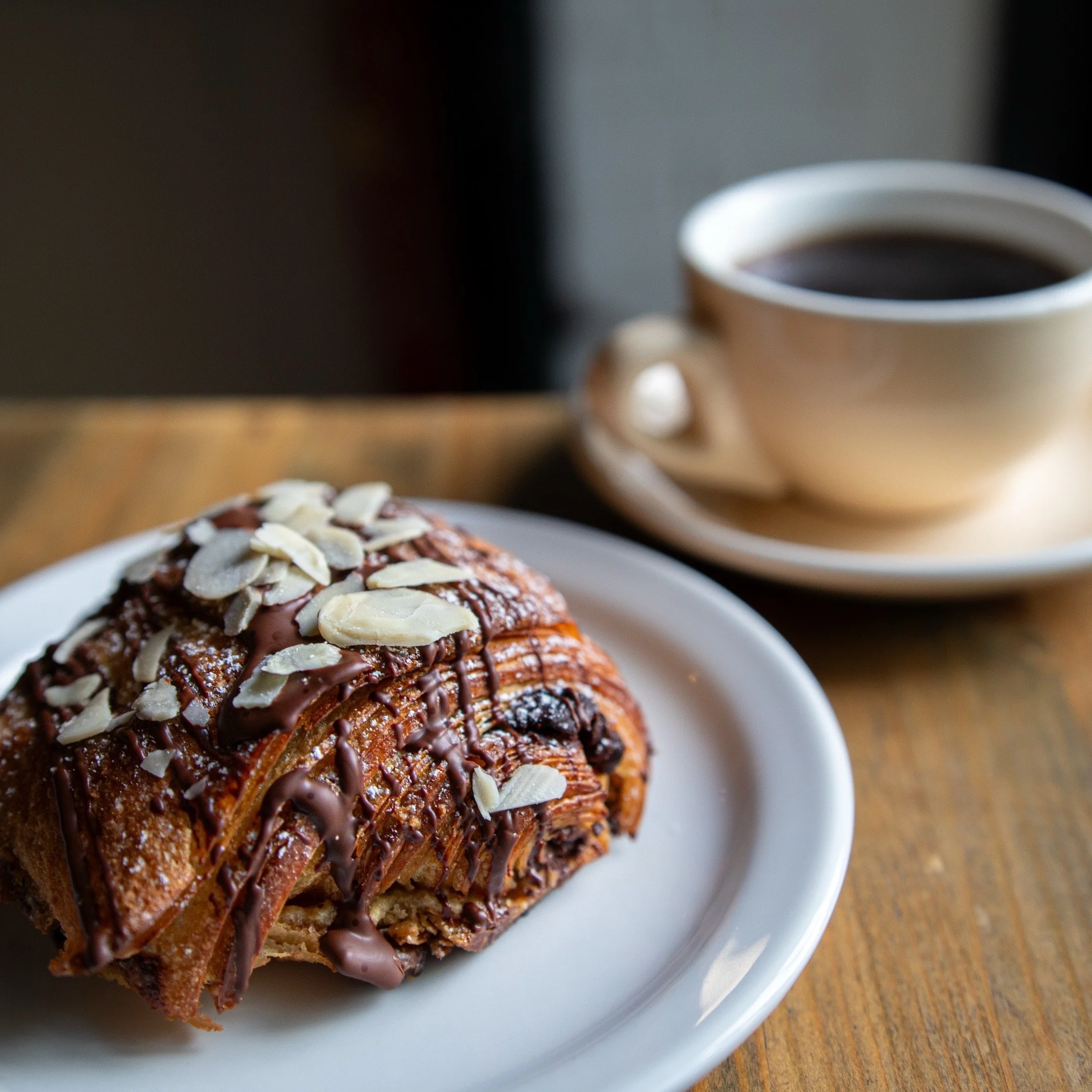 A croissant drizzled with chocolate sauce and topped with sliced almonds on a white plate.