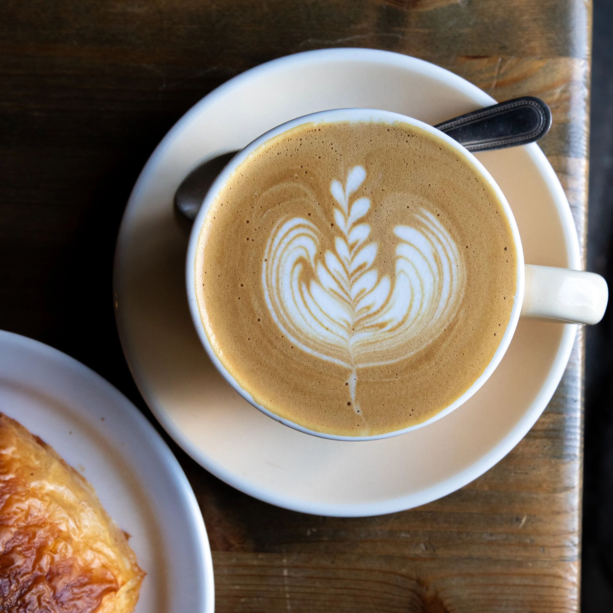 A close-up view of a cup of coffee with latte art on top, placed on a white saucer, with a spoon beside it, on a wooden table, partially showing a croissant on a white plate.