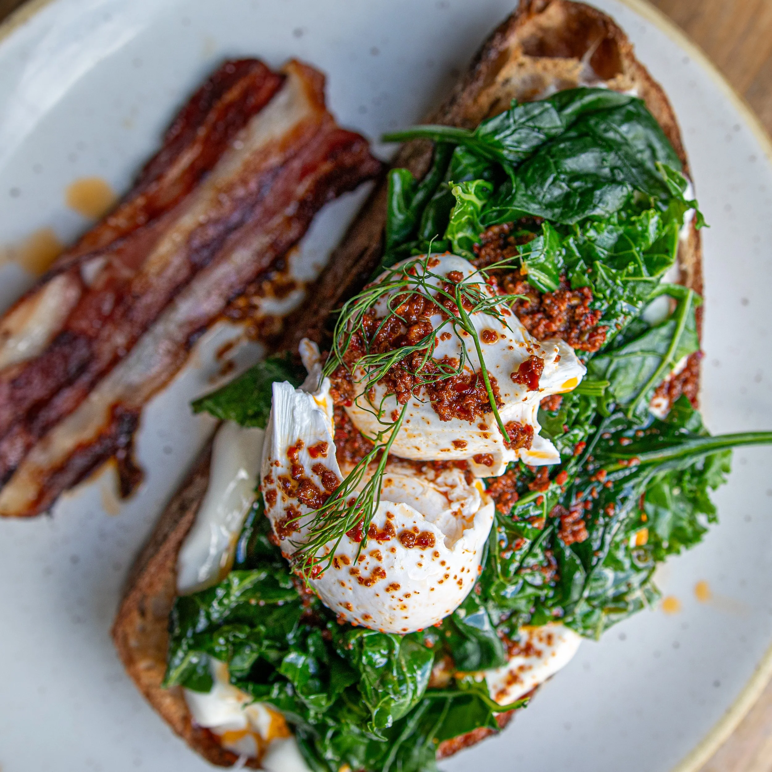 A presentation of a breakfast dish with toast, greens, soft-boiled eggs, tomato-based sauce, and crispy bacon on a white plate.