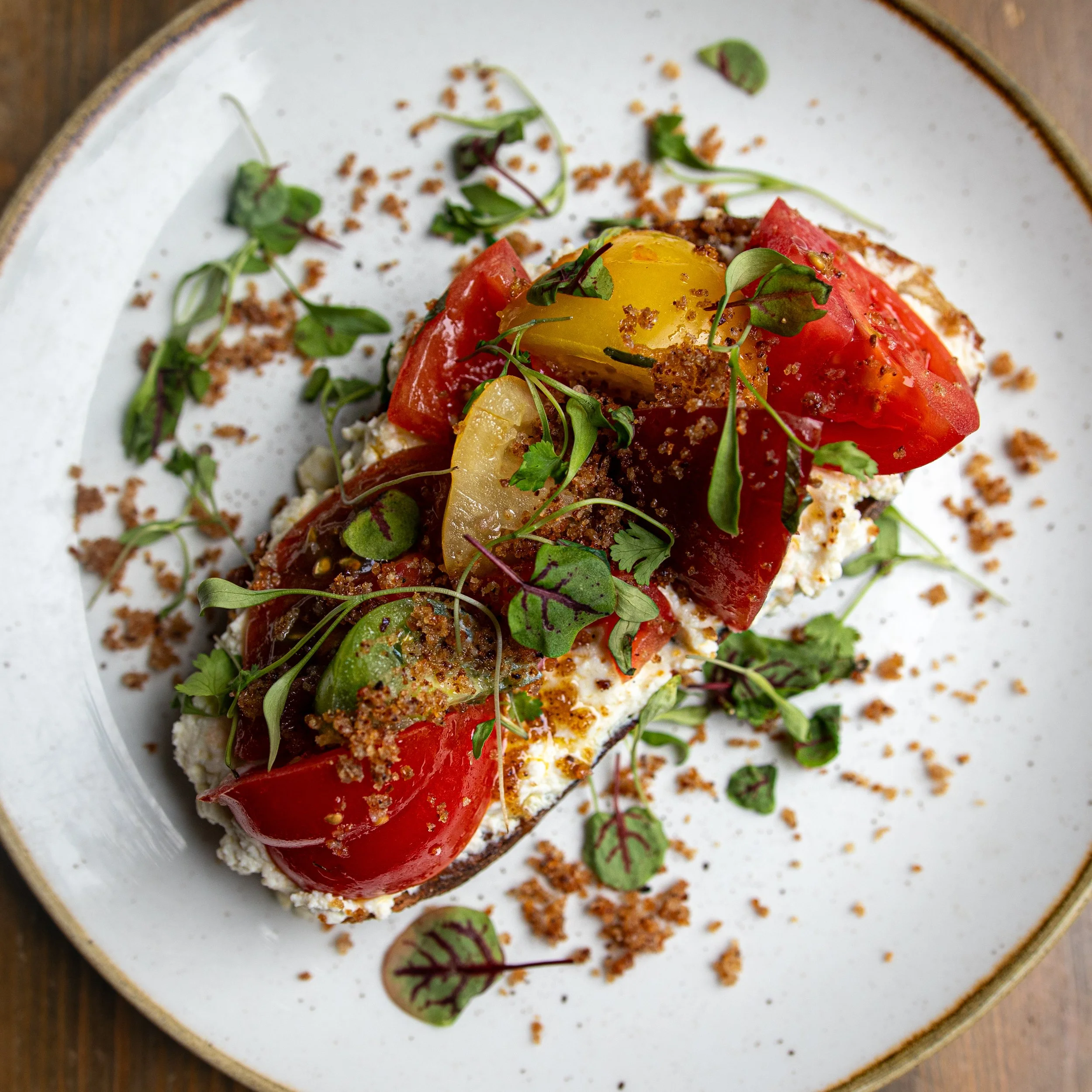 Close-up of a gourmet dish featuring roasted red peppers, a soft-cooked egg on top, microgreens, and crumbled toppings on a white plate with a thin rim.