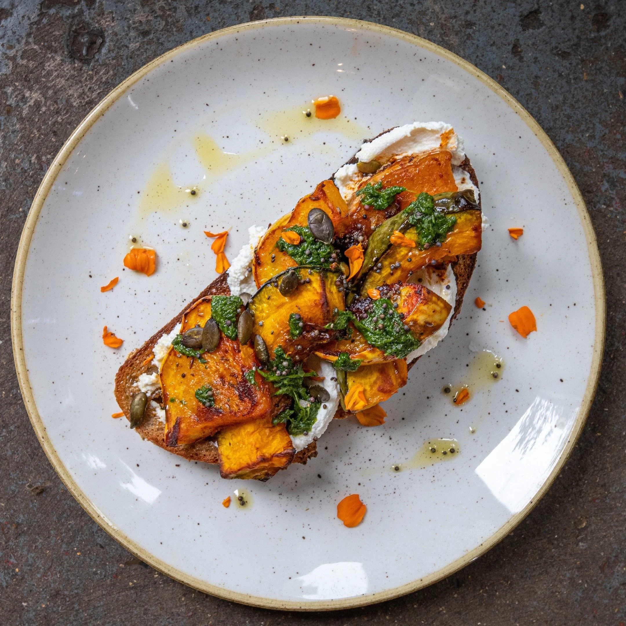 A slice of squash toast topped with herbs, seeds, and cheese on a white ceramic plate with scattered flower petals and peppercorns, placed on a dark textured surface.