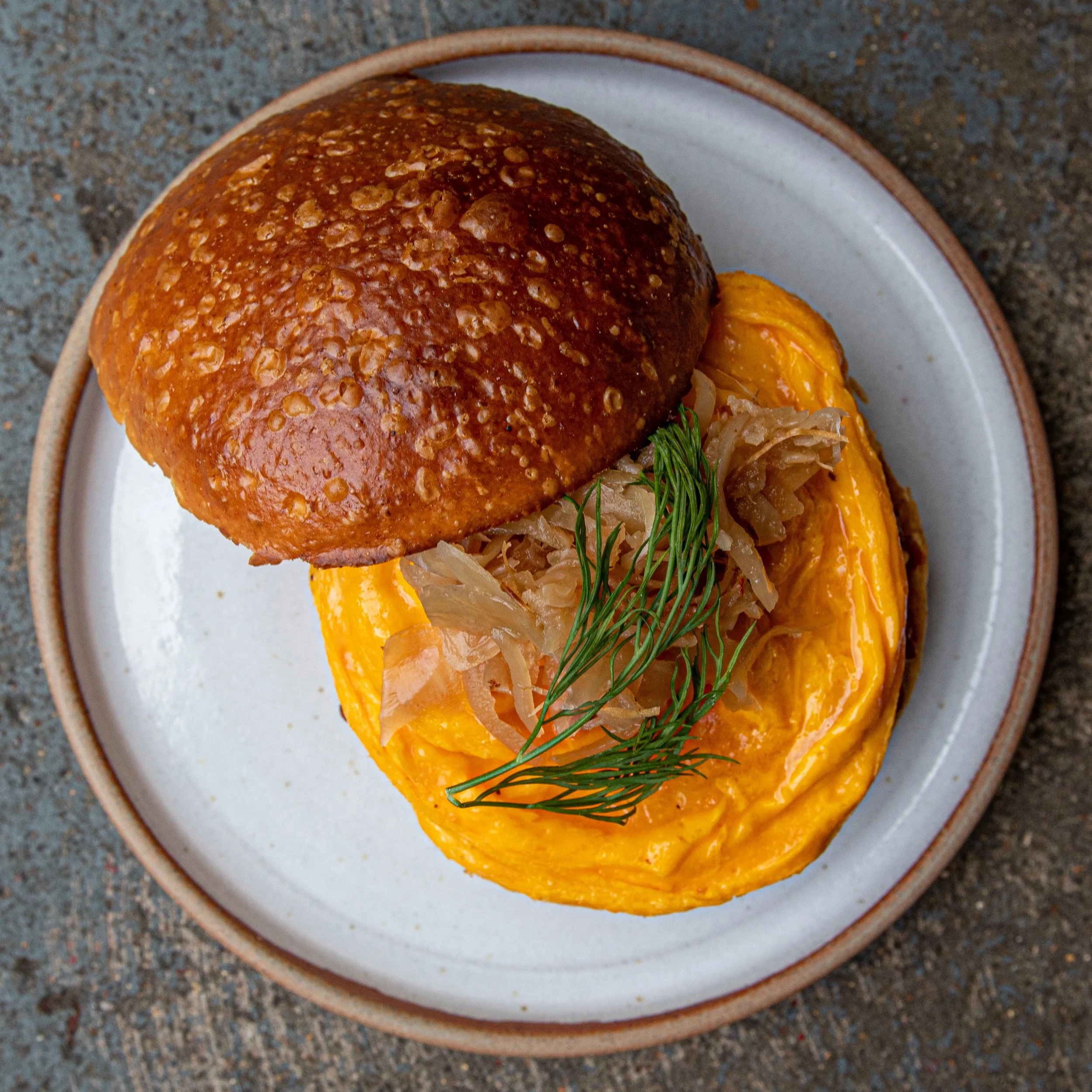 A close-up of a bread roll with sesame seeds on top, served on a white plate with a brown rim, placed on a textured dark surface.