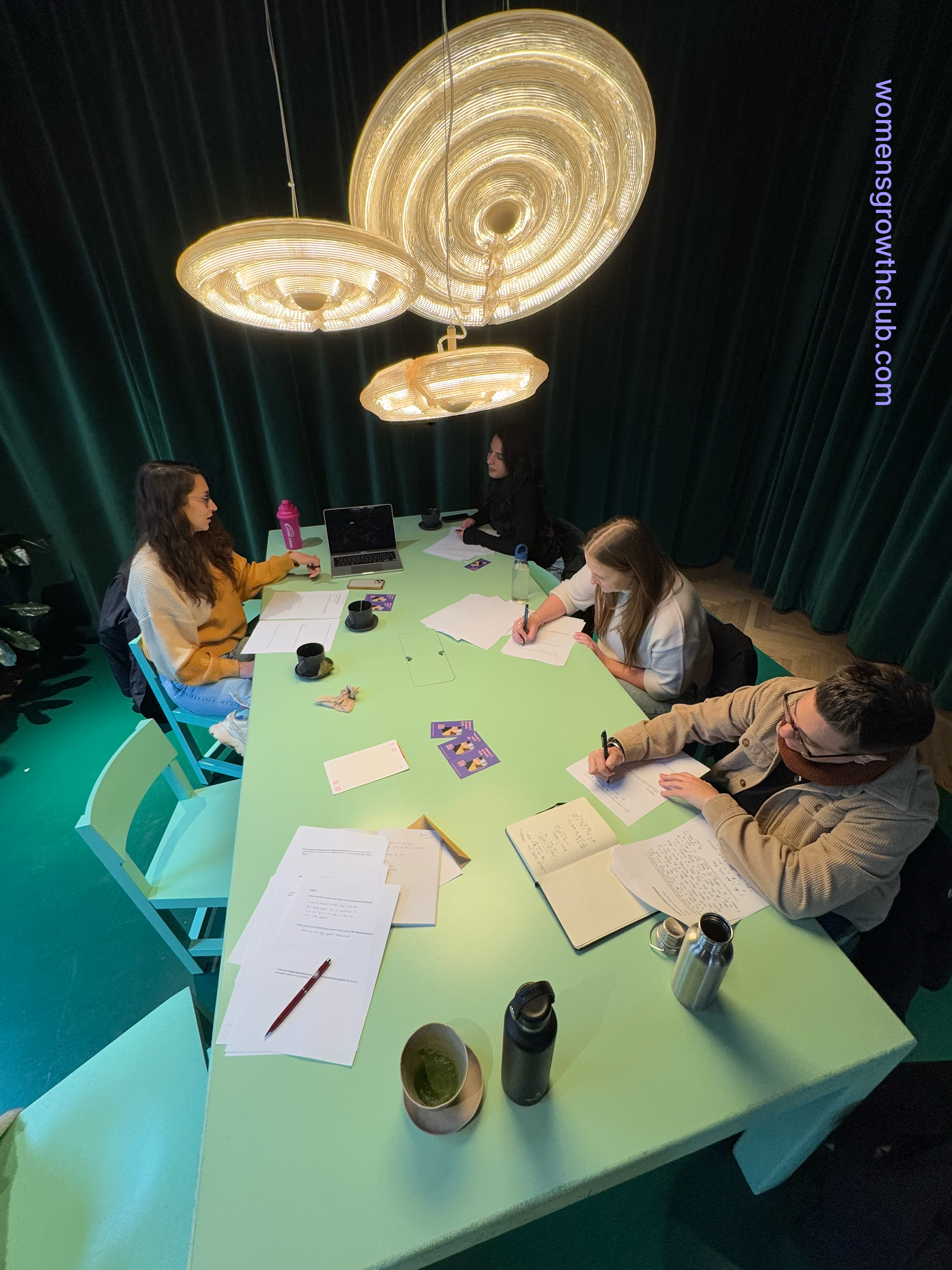 Four women sitting around a green table in a dimly lit room with three large, ornate chandeliers hanging above. The table has various papers, notebooks, pens, cups, water bottles, and a laptop.
